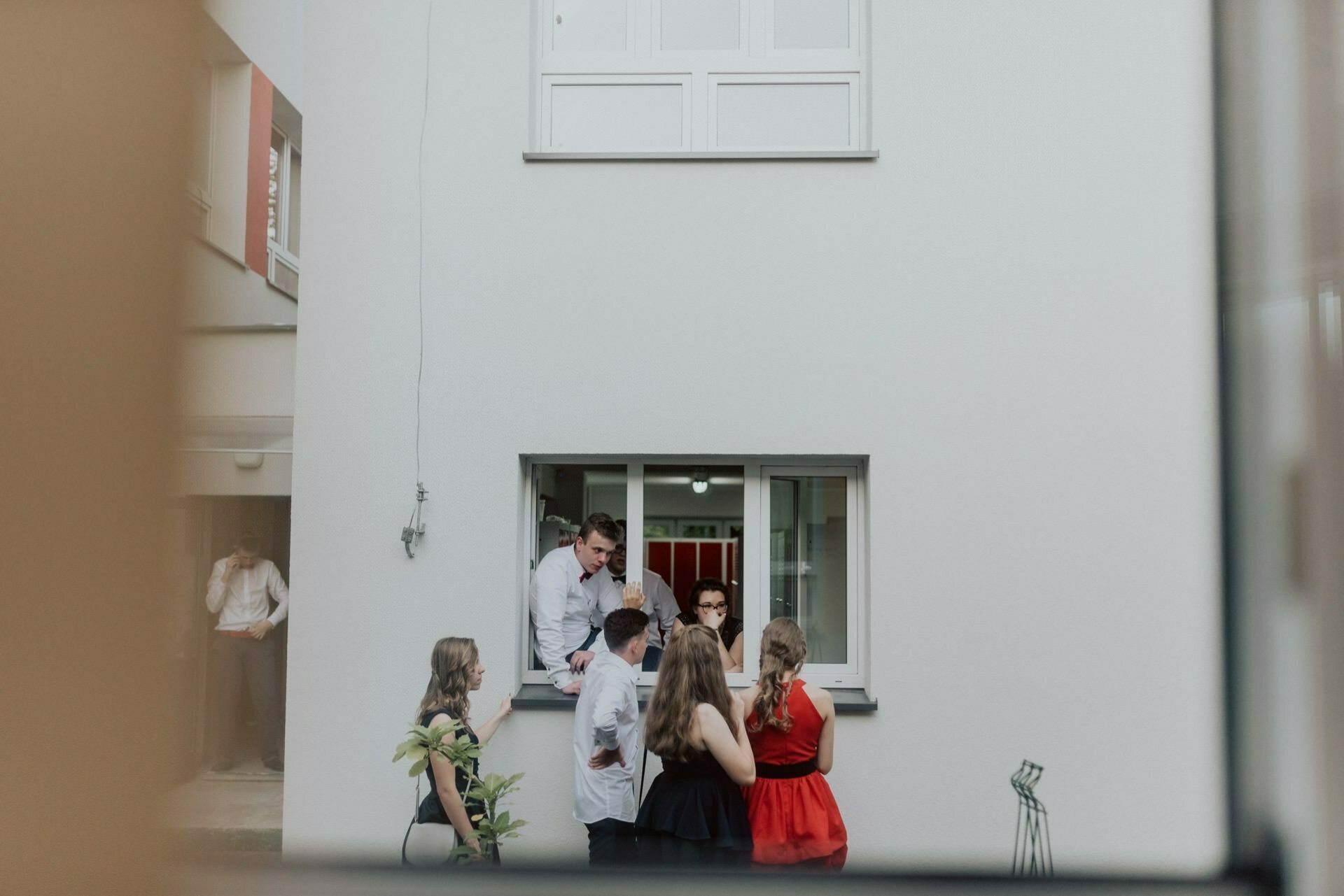 A group of people gather in front of a white building, captured in a stunning event photograph. Some, seen through an open window, are leaning over and talking to someone inside. One person, dressed in semi-formal attire consisting of white shirts and dresses, stands alone and talks on the phone.  