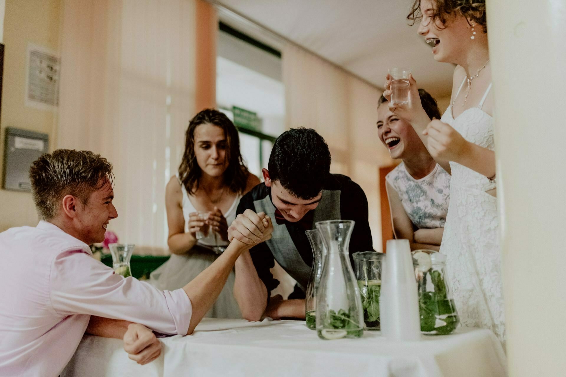 Five people gather around a table and play a friendly arm wrestling match. Two people take part in the competition, while three others watch and cheer with smiles and laughter. Pitchers and cups are placed on the table, creating a fun and relaxed scene, perfect for any photo essay of the events.  