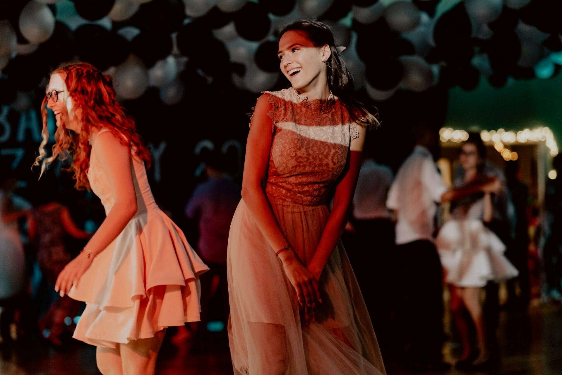 Two young women in dresses dance joyfully under a canopy of balloons at a party. One wears glasses and a short pink dress, while the other wears a long lace gown. The background, perfectly captured by our event photographer Warsaw, is filled with other partygoers and low lights.  