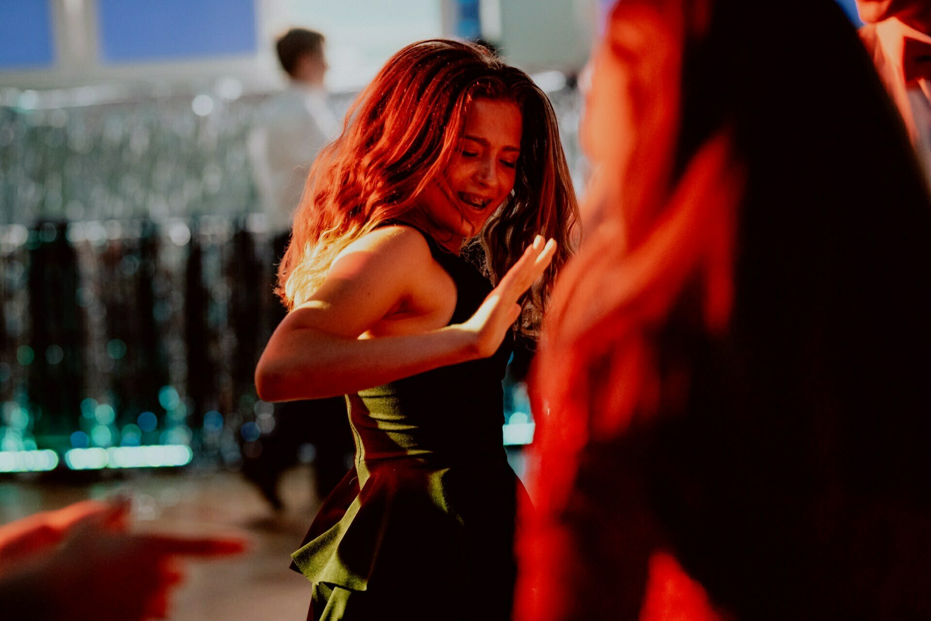 A girl with long, wavy hair, wearing a black dress, dances vigorously with her eyes closed and hands raised. The background is blurred and the vivid red and blue lighting creates a lively atmosphere. Other dancing figures are partially visible in this stunning event photograph.  