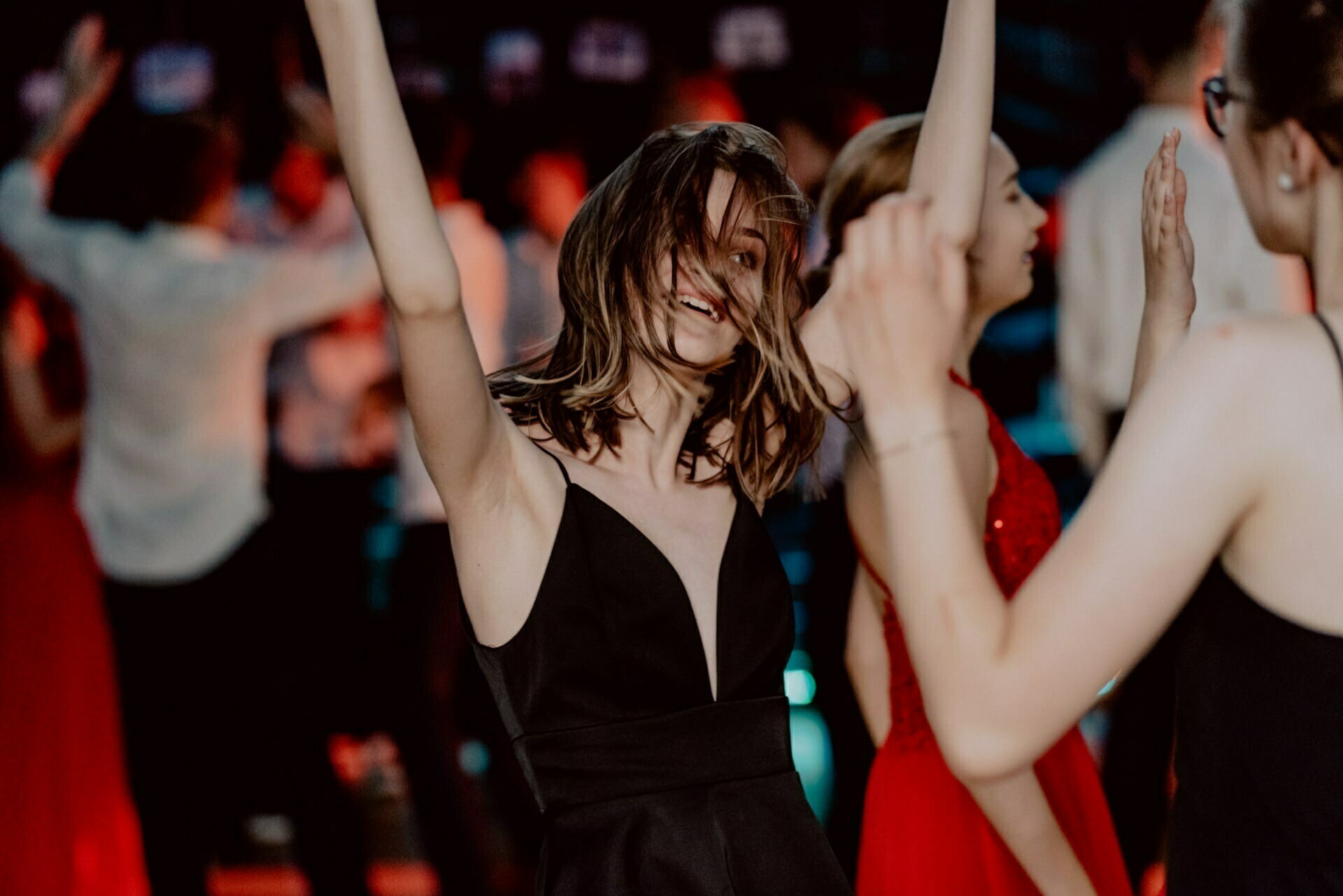 A young woman in a black dress dances with her hands raised and her hair partially covering her face during a festive event captured in a captivating photo essay of the events. Other people around her, some in red dresses, are also dancing and having fun. The background is blurred with colorful lights.  