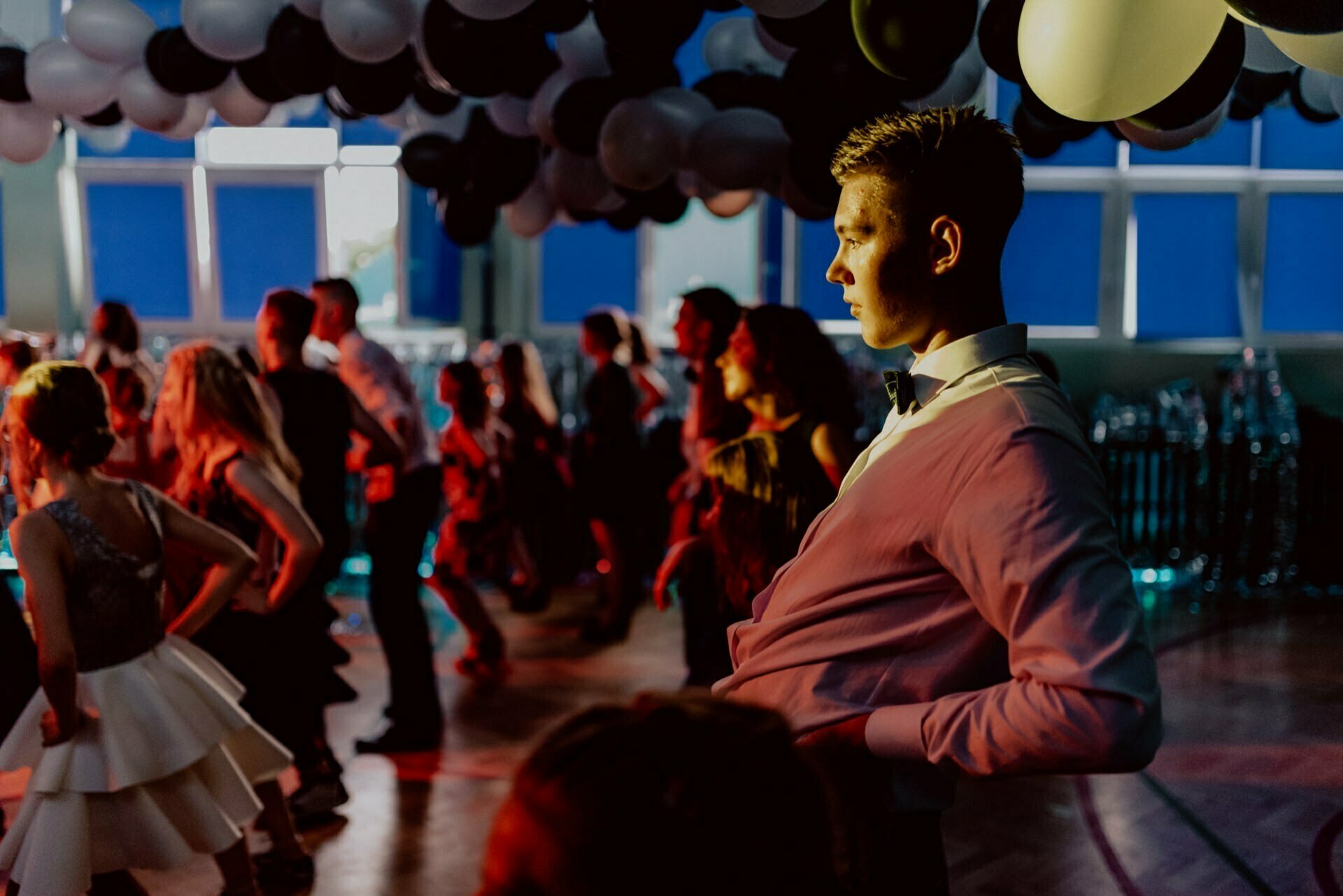 A young man in formal attire dances under a ceiling filled with black and white balloons illuminated by a mix of colorful lights. In a dimly lit room with large windows, several other people can be seen dancing, and a photographer for the junior high school prom has captured this vibrant scene. 