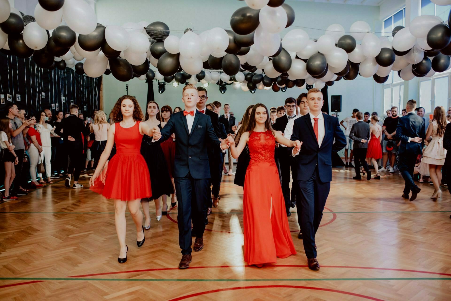 A group of young people dressed formally in red and black outfits dance in a decorated hall. Black and white balloons hang from the ceiling. Additional people, standing in groups against the walls, watch from the background. This scene perfectly captures the vibrant photo-op of the events.   