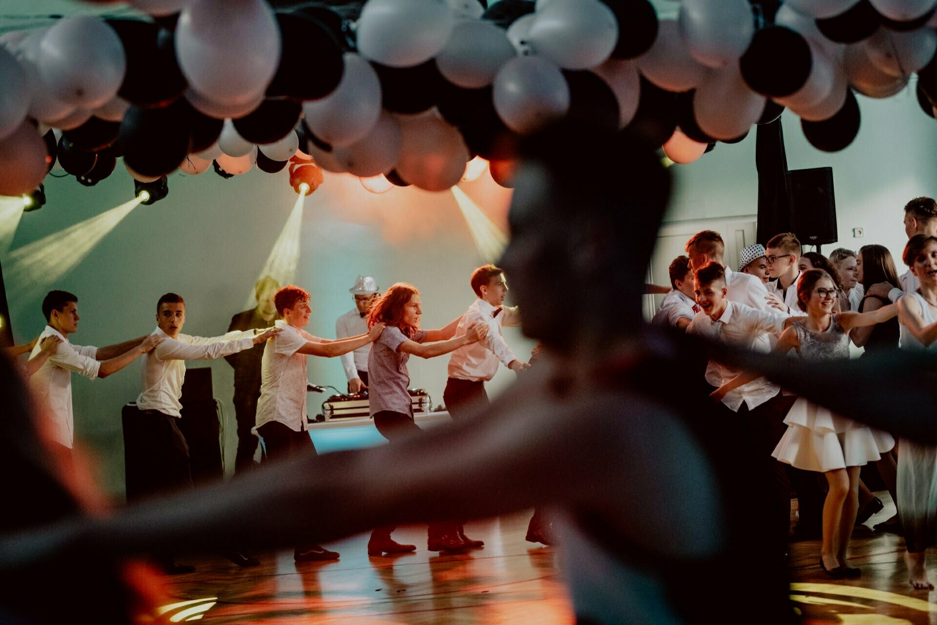 A lively dance party with people forming a conga under a ceiling decorated with black and white balloons, perfectly captured by an event photographer Warsaw. Multicolored lights illuminate the room, and a DJ can be seen in the background. The event photo report shows how the guests thoroughly enjoyed the celebration.  