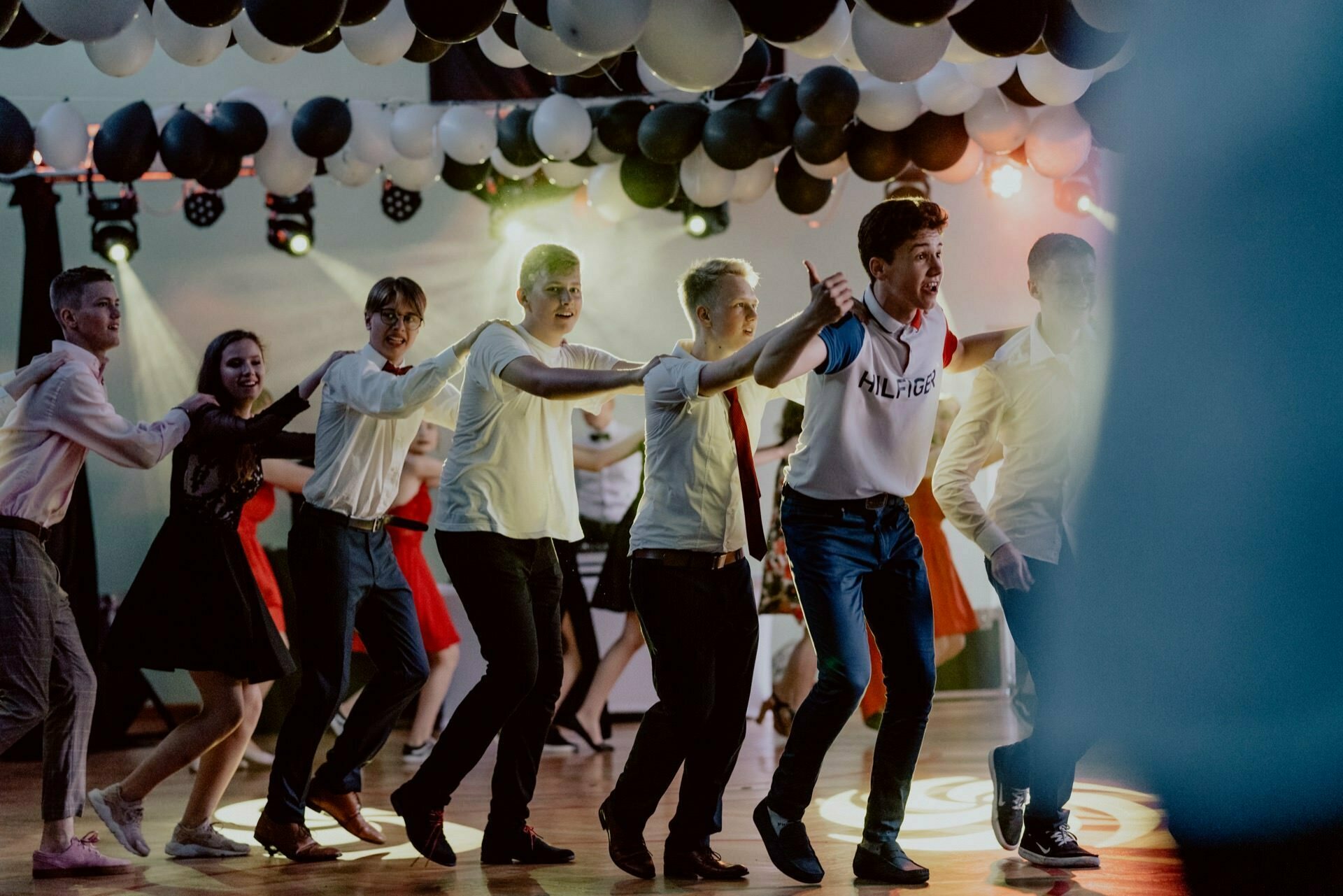 A group of people merrily dancing in a conga row under black and white balloons is captured in this vibrant photo essay of the event. They are dressed in a mix of formal and casual attire, enjoying the lively atmosphere as multicolored lights illuminate the dance floor. 