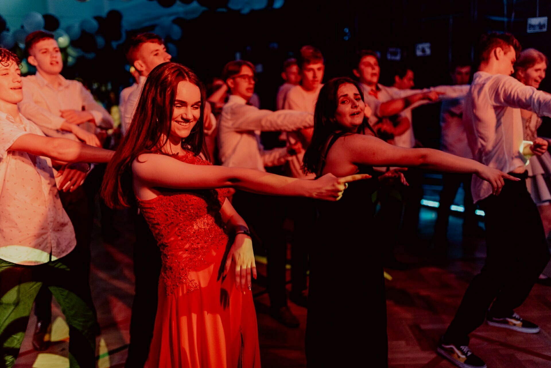 A group of young people are dancing in a dimly lit room with colored lights. The centerpiece is a young woman in a red dress, joyfully dancing with others around her. The lively and festive atmosphere suggests celebration, beautifully captured in event photography by a talented event photographer Warsaw.  