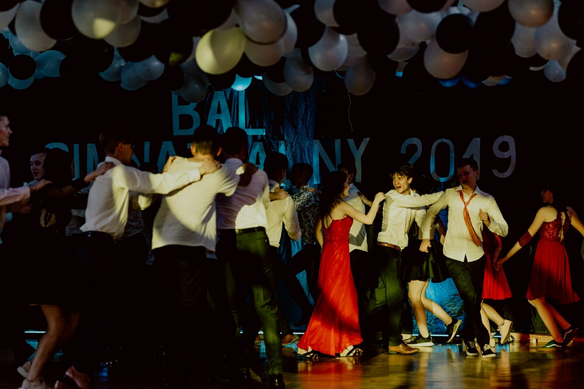 A group of people dressed in formal attire dance energetically in a dimly lit room decorated with black and white balloons. In the background, the year "2019" and the word "Ball" are visible, suggesting a celebratory event, which was perfectly captured in an event photo report by a talented event photographer Warsaw. 