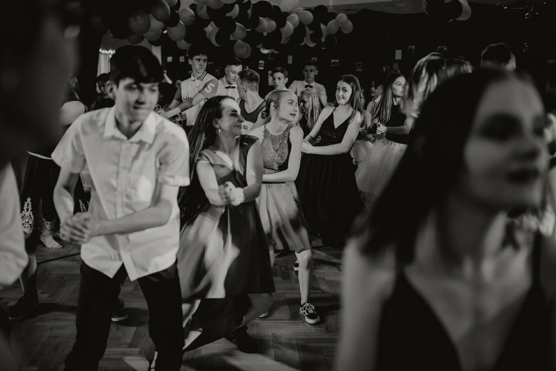 A large group of young people dance close together in a dimly lit room decorated with hanging balloons and streamers. Some wear formal attire, while others are more casually dressed. The atmosphere seems lively and festive, perfectly capturing the essence of event photography by event photographer Warsaw.  