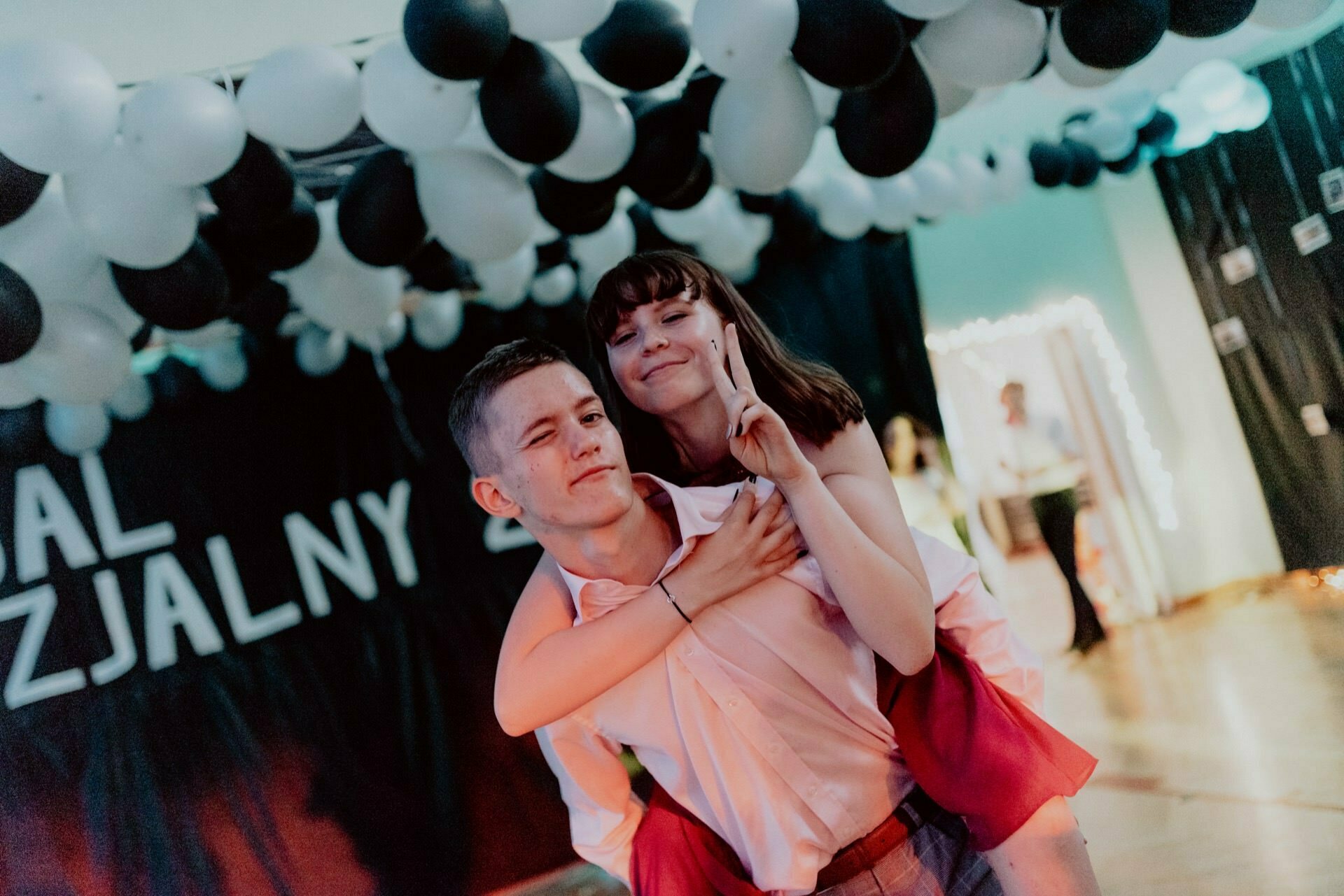 A young man in a light-colored shirt carries a smiling young woman on his shoulder, who makes a peace sign with his fingers. They are under a ceiling decorated with black and white balloons and a black draped background with partially visible text - a perfect snapshot of the events. 