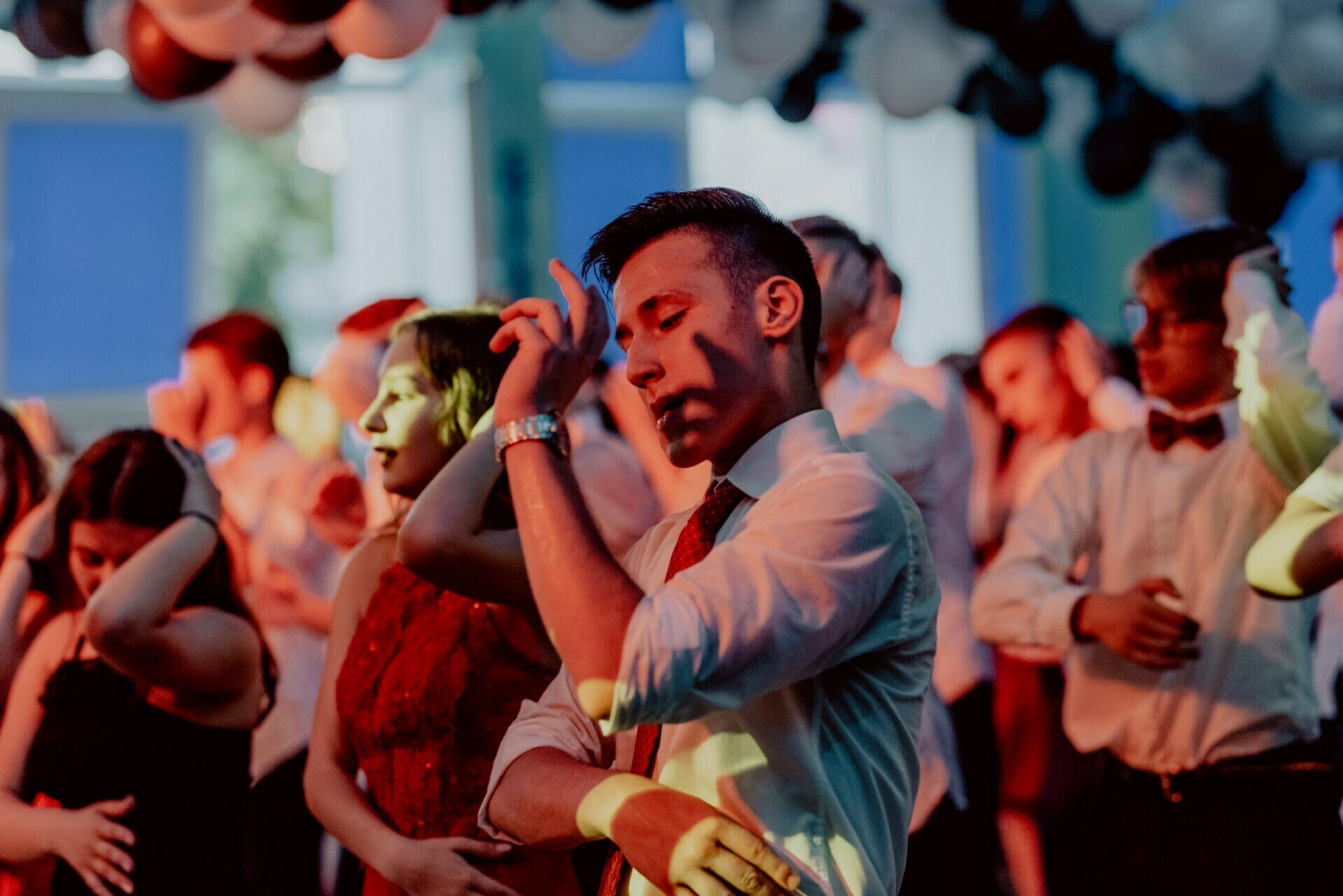 A group of people dance under a ceiling decorated with balloons of different colors. Individuals are dressed formally, with men in shirts and bow ties and some women in elegant gowns. The colorful lighting creates a festive atmosphere as they move in sync, which was beautifully captured by event photography.  