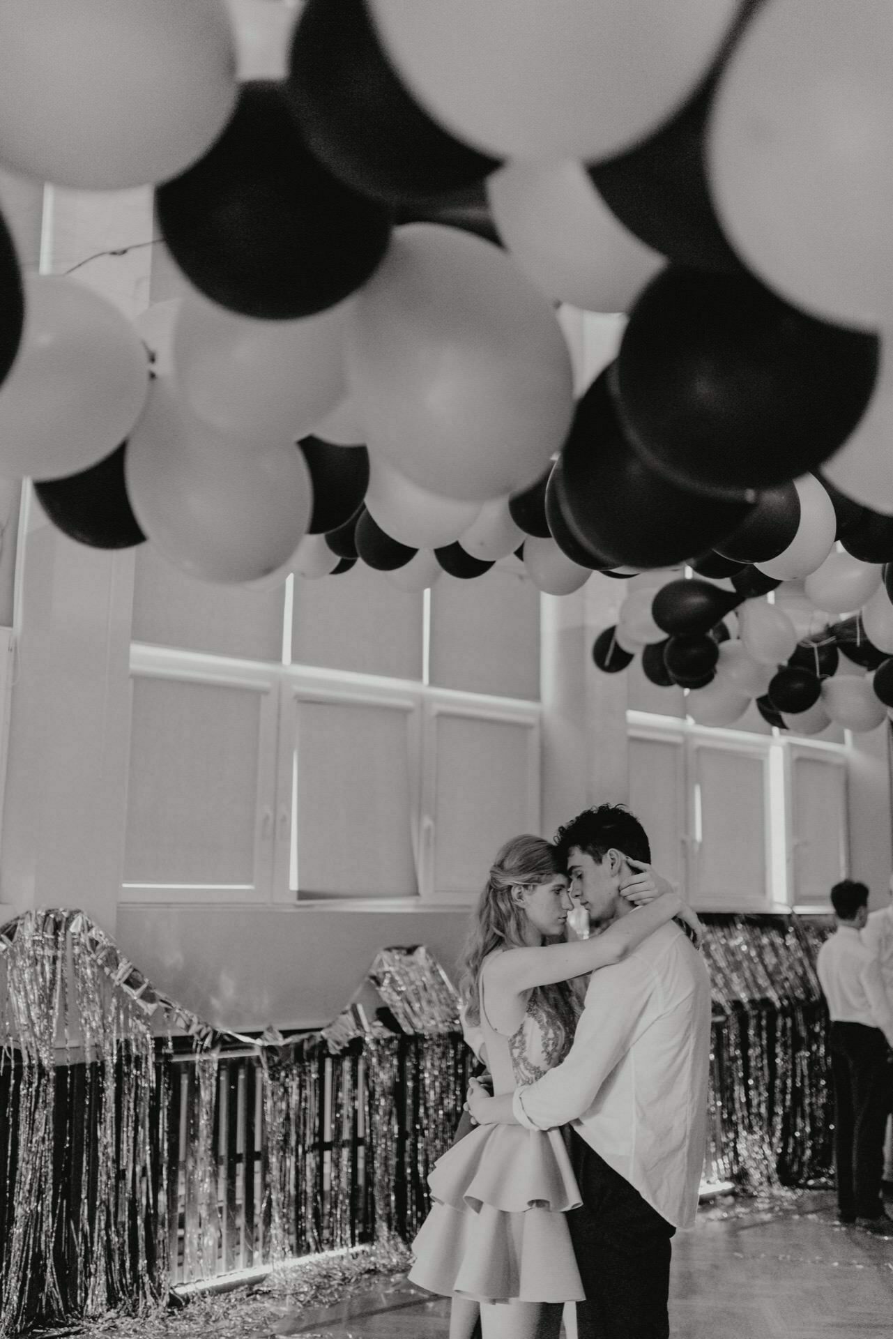 Black and white photo showing a couple embracing and dancing under a ceiling decorated with black and white balloons. The couple is surrounded by glittering decorations hanging from the ceiling, creating a festive atmosphere. This captivating event photography captures the joy of celebration.  