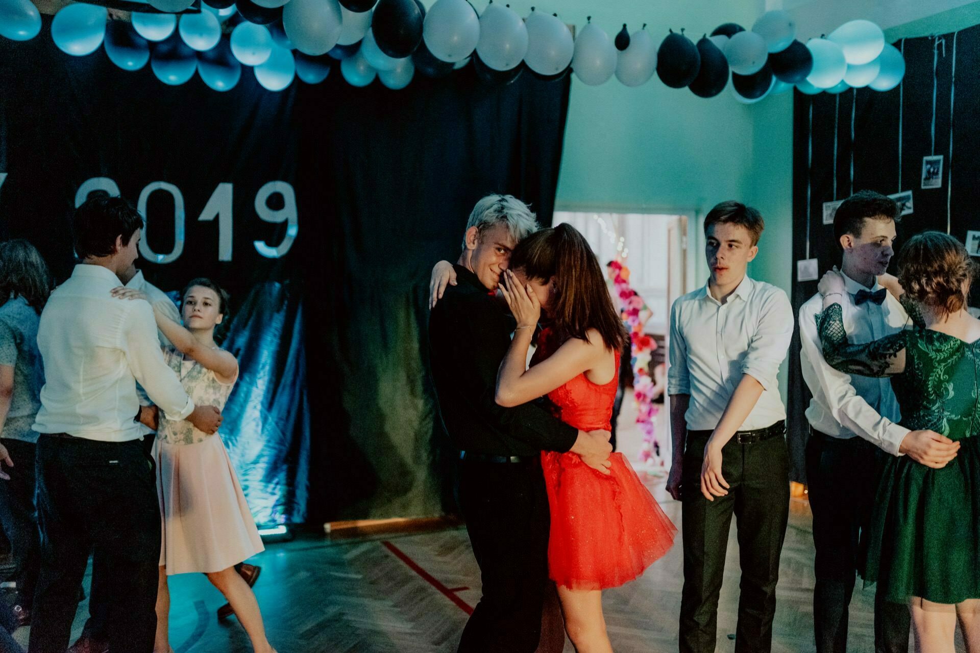 A group of young people dance during a decorated party with balloons on the ceiling and "2019" written on the wall. In the middle, a couple slowly dances close; a girl in a red dress covers her face with one hand. Others are dancing and talking - a perfect scene for event photography.  