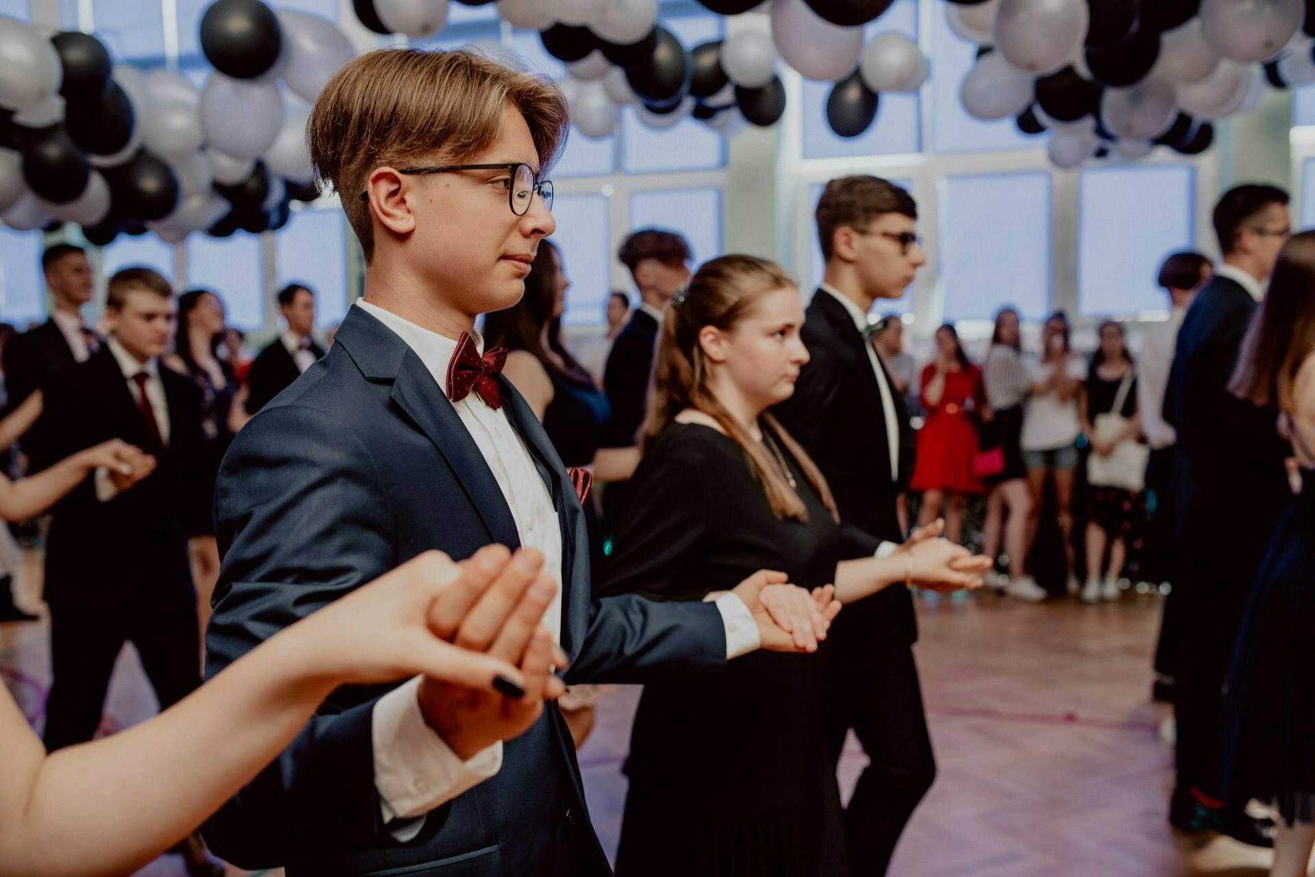 A group of young people dressed in formal attire are dancing in a ballroom decorated with black and white balloons. One couple, a young man in a blue suit and glasses and a young woman in a black dress, hold hands and focus on their dance steps. The audience watches, perfectly capturing the essence of event photography.  