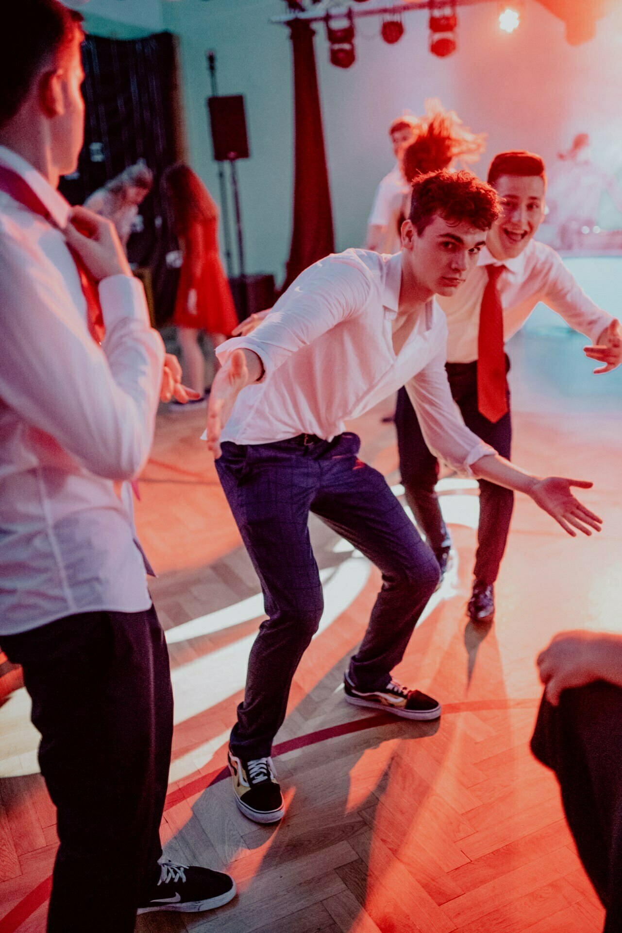 A group of young men dressed in white shirts and dark pants dance vigorously at a lively party. One of them adopts a crouched, playful pose, while another standing behind him enthusiastically points toward the camera. The backdrop is illuminated with red and pink lights - perfect for event photography in which to capture the moment.  