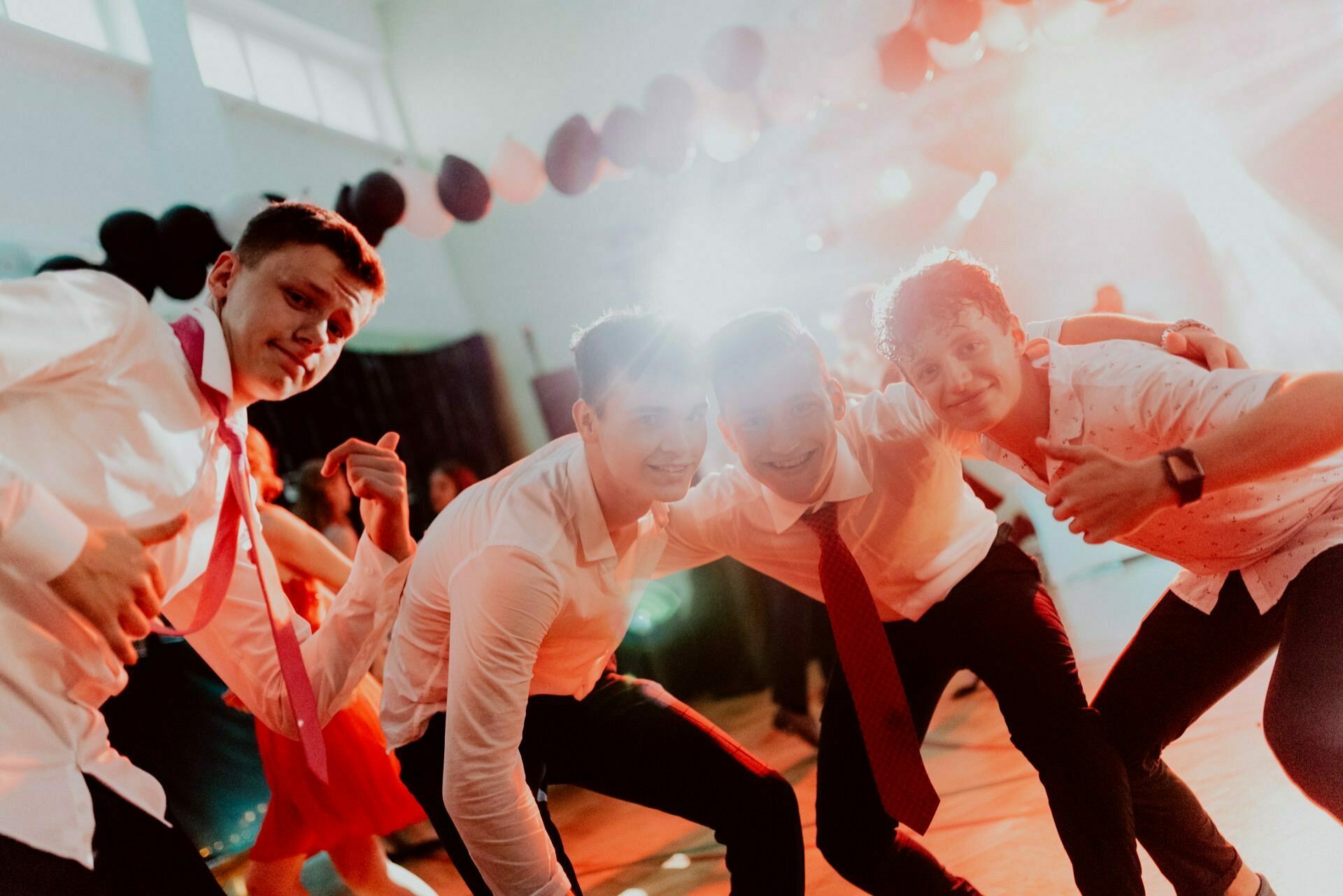 Four young men, dressed in white shirts and ties, smile and pose for the camera in a festive, well-lit room. Black and red balloons are visible in the background, suggesting a festive event. One person raises his thumb in the air, capturing a perfect photo-op of the events.  