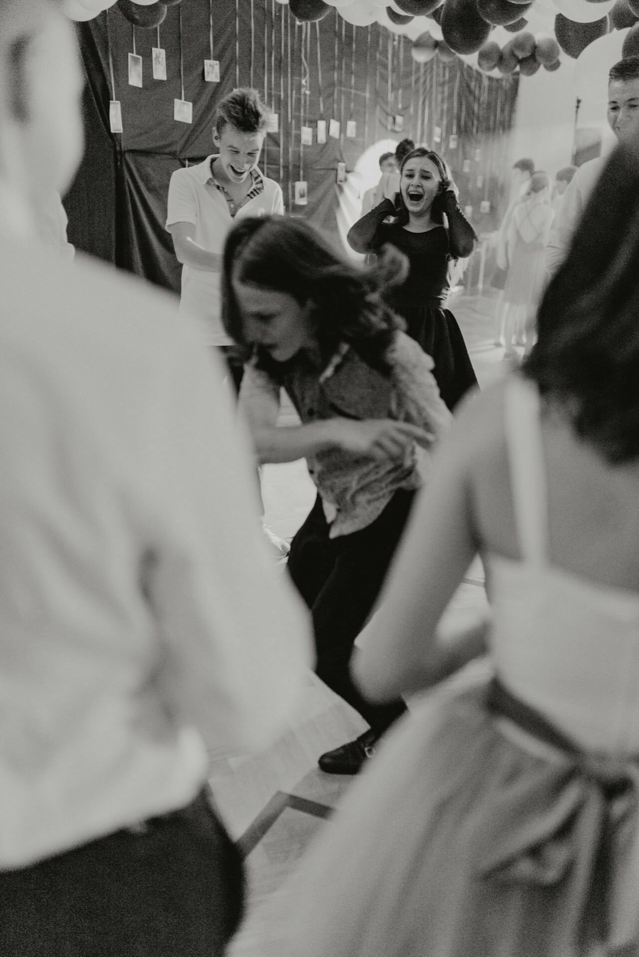 The black-and-white photo shows a vibrant scene at a party. In the foreground, someone is dancing energetically, while others are watching and smiling. Balloons and decorations hang from the ceiling, adding to the festive atmosphere - presenting event photography at its finest.  