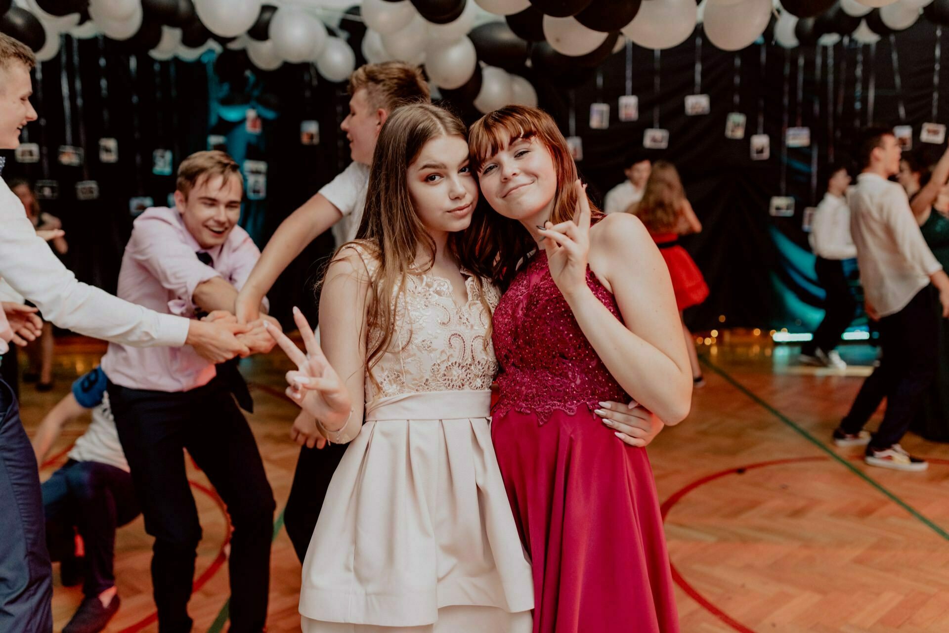 Two young women pose for a photo on a decorated dance floor. One is wearing a white dress, the other a red one. They both give a peace sign and smile. There are people dancing in the background, and the ceiling is decorated with balloons with hanging Polaroid photos, capturing the essence of event photography.   