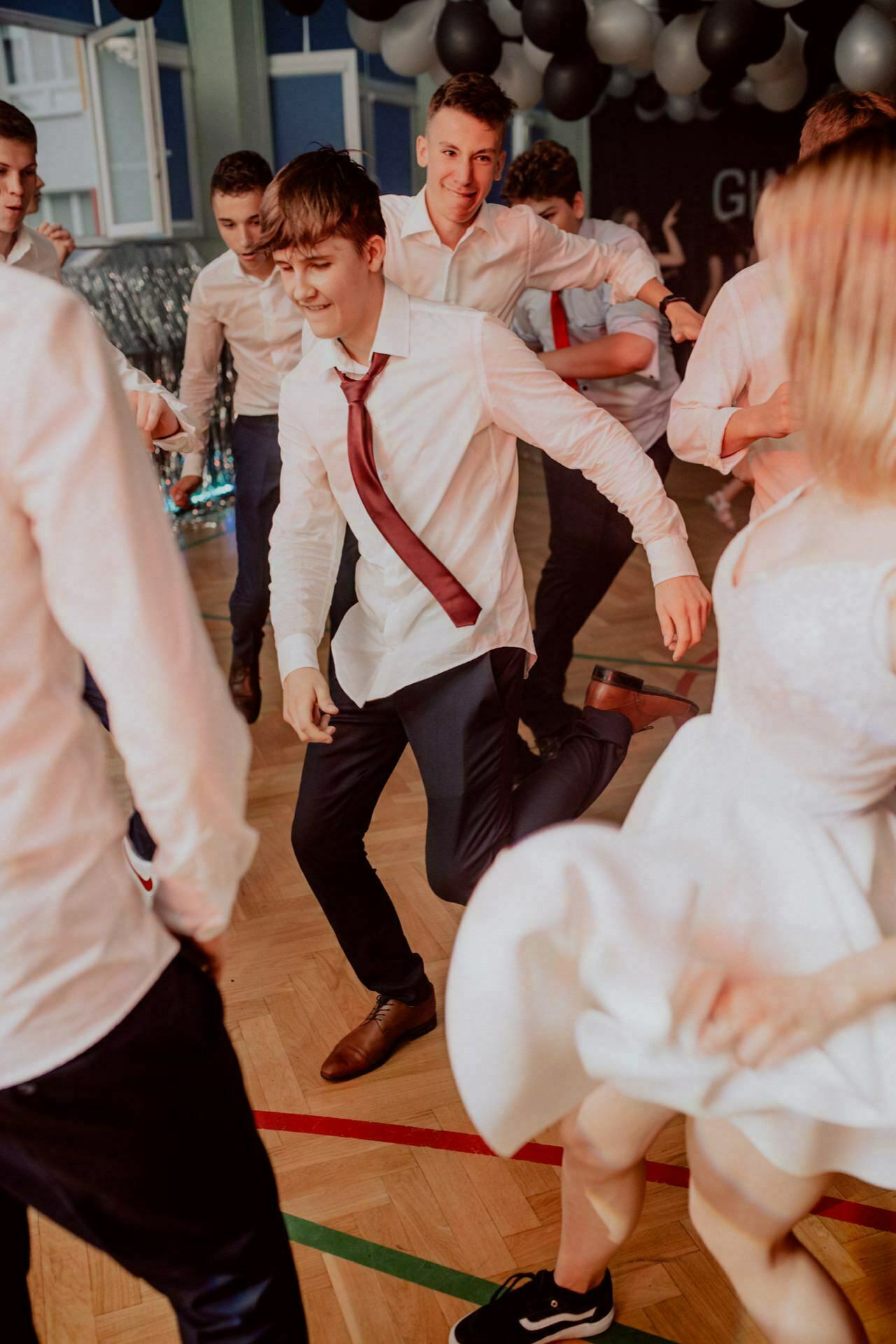 A group of young people in formal attire dance vigorously during a ceremony. The central figure, a young man in a white shirt and red tie, is in mid-motion and clearly enjoying himself. Balloons and festive decorations are visible in the background, perfectly captured by event photographer warsaw in a delightful event photo essay.  