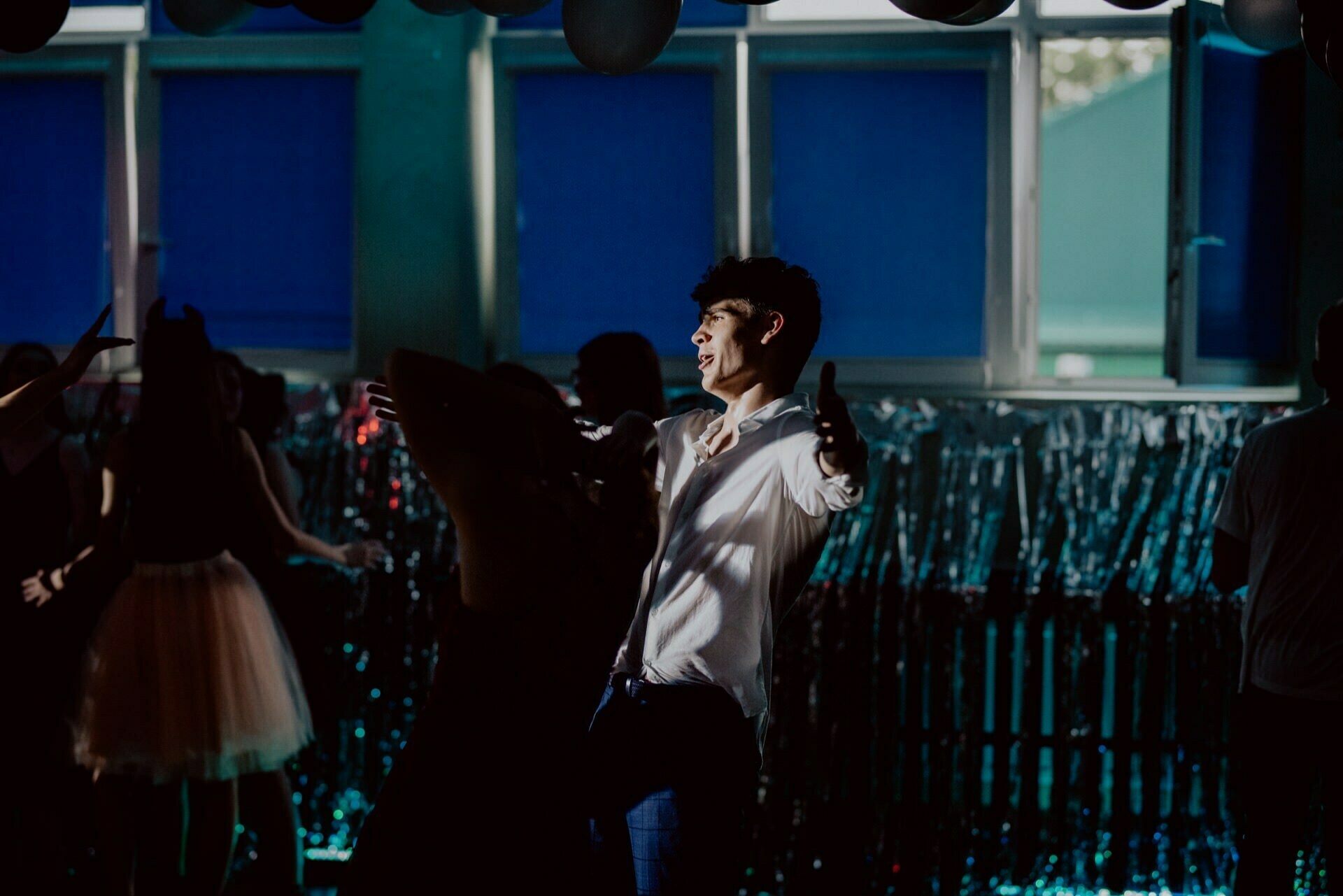 A person wearing a white shirt dances in a room decorated with streamers and balloons. In a dimly lit background, dancing people can be seen, with blue windows visible behind them. This scene captures the essence of a vibrant photo-relation of the events.  