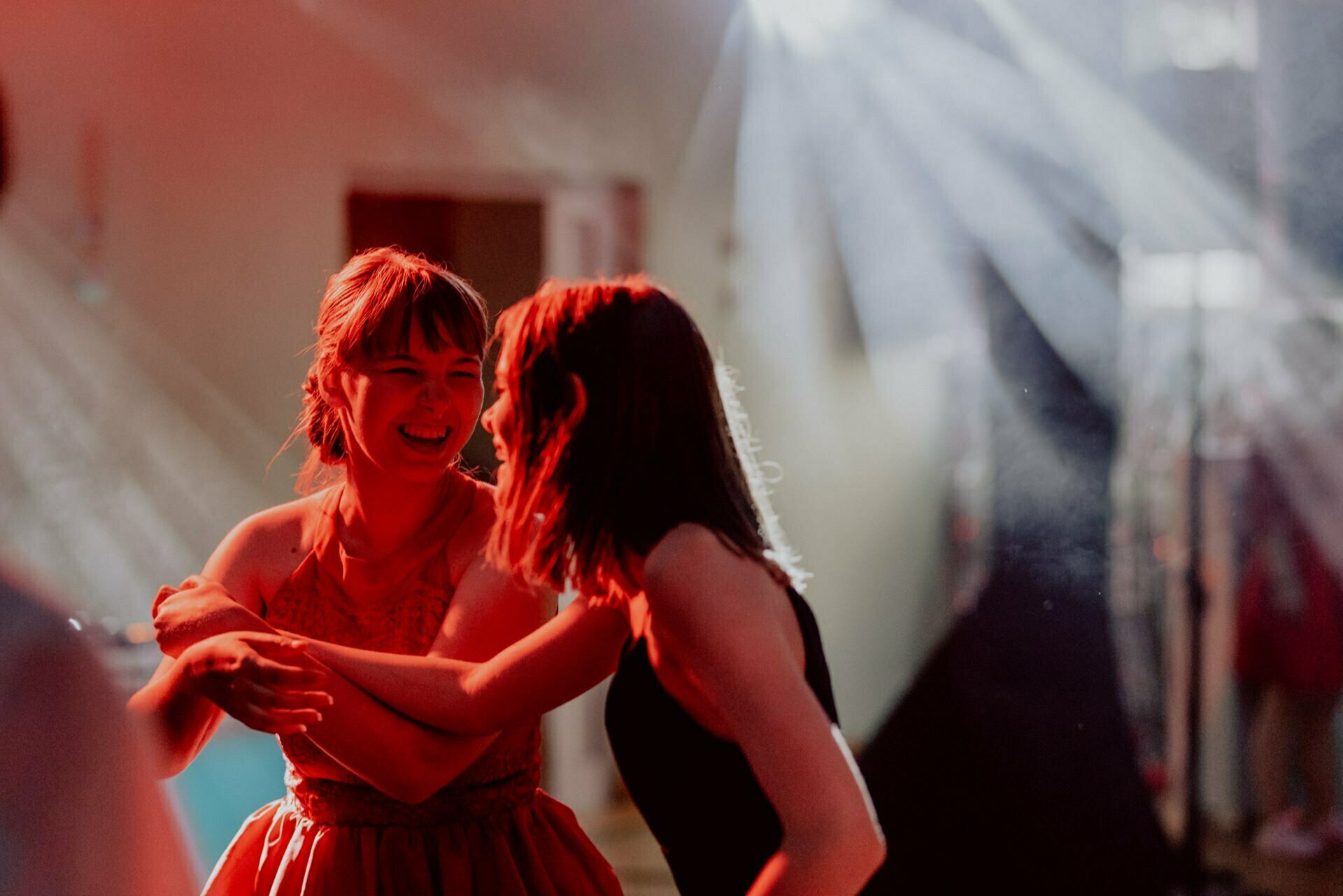 Two women joyfully dance together at a party illuminated by dynamic red and white lighting. They seem to be in motion, with one woman embracing the other with her arm. The background is blurred, emphasizing the vibrant atmosphere of the event - exquisite event photography capturing pure happiness.  