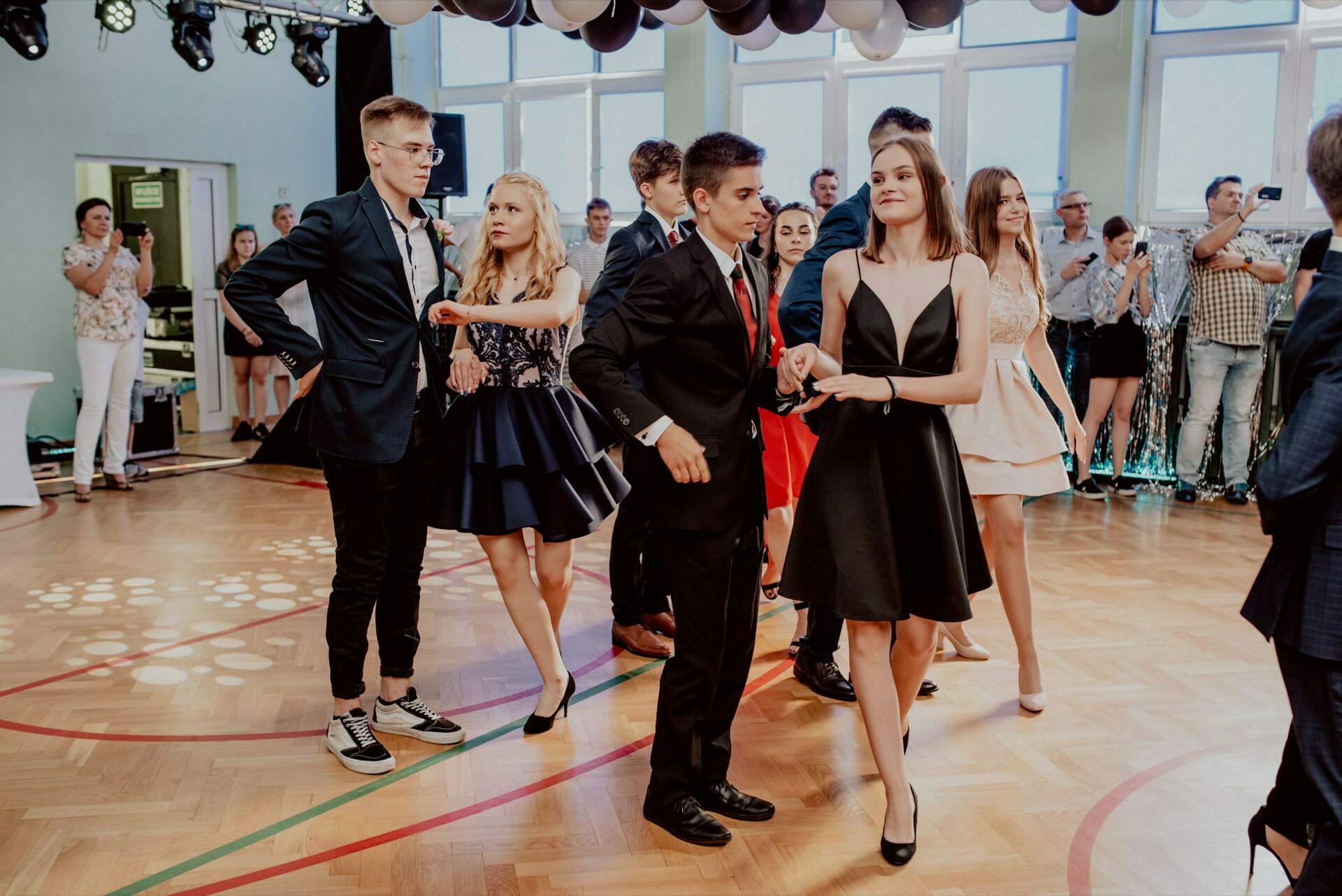 A group of young people dressed in formal attire dance in a brightly lit room with balloons on the ceiling. Two pairs of dancers are visible in the foreground. In the background are spectators and photographers, including an event photographer Warsaw, capturing a snapshot of the events.  