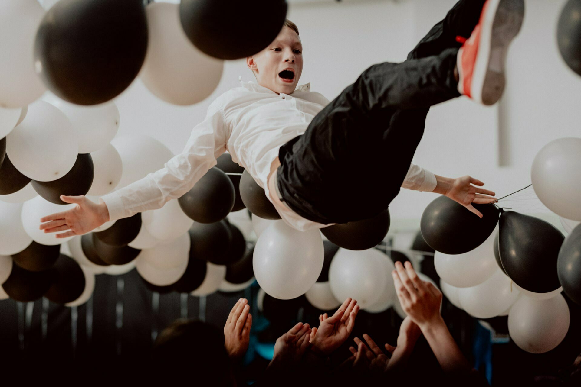 A person wearing a white shirt and black pants is tossed into the air by a group of people, with his hands visible below. The background is decorated with black and white balloons. The person seems to be enjoying the moment with an excited expression on his face, which is perfectly captured in this vibrant event photography.  