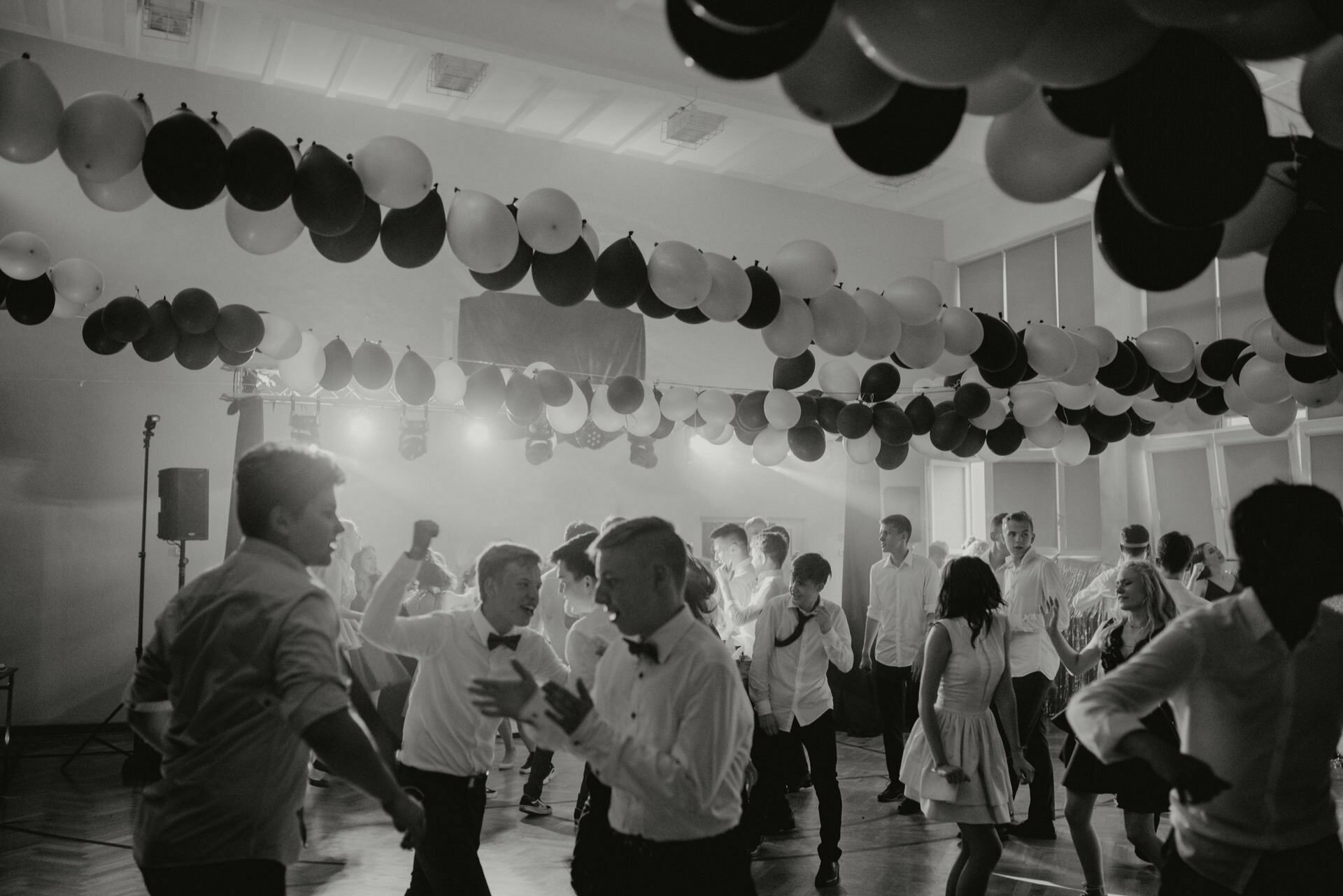 A lively dance floor in a decorated room with balloons hanging from the ceiling. Men in formal attire and women in dresses dance vigorously under bright lights, while a DJ can be seen in the background. The photo capturing this lively moment of event photography is in black and white.  