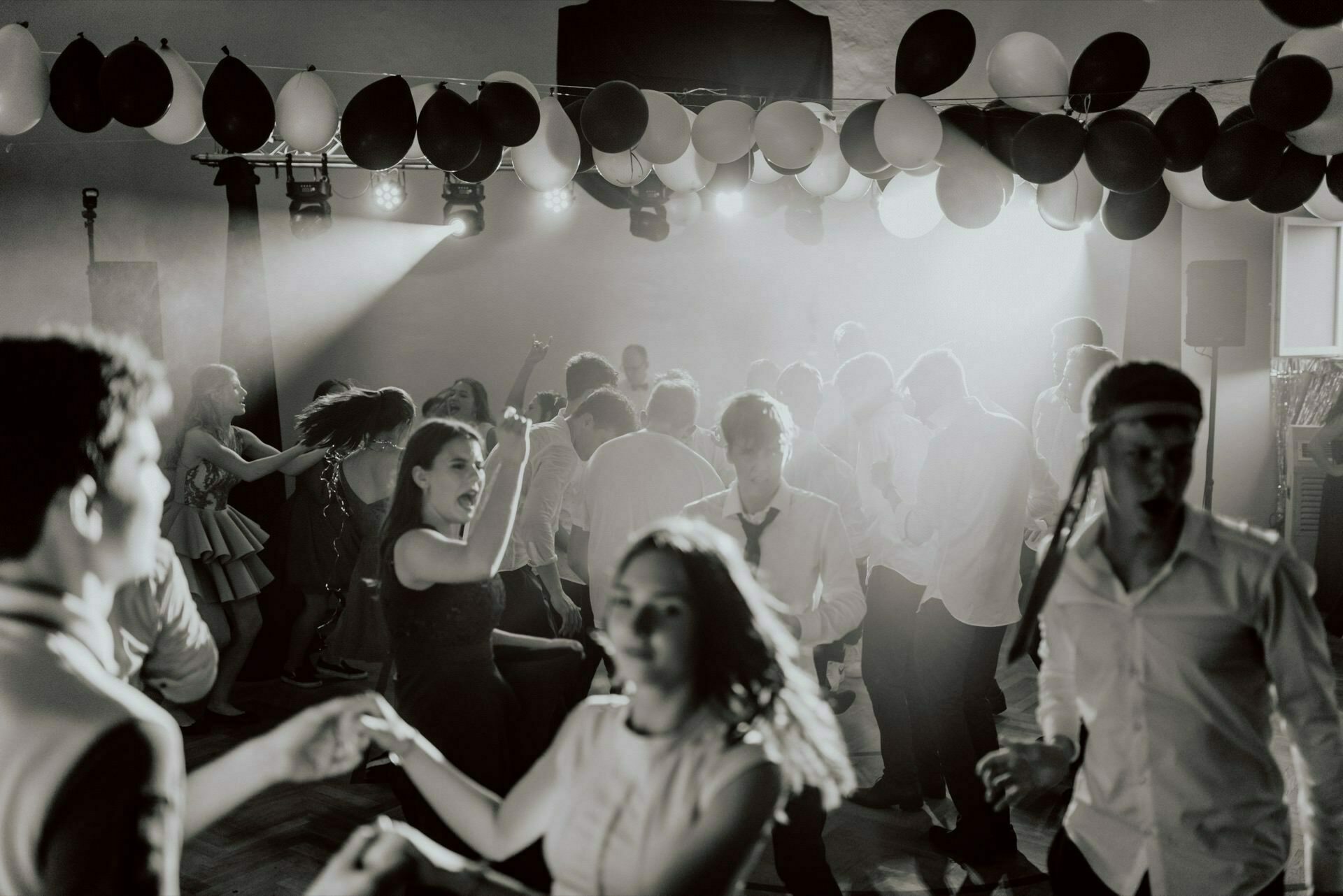 A group of people dancing energetically at a party, beautifully captured in event photography. The room is decorated with lots of balloons, and spotlights cast a dramatic light on the space. Most of the attendees are dressed in semi-formal attire, and many are dancing in pairs or groups. Black and white image.   