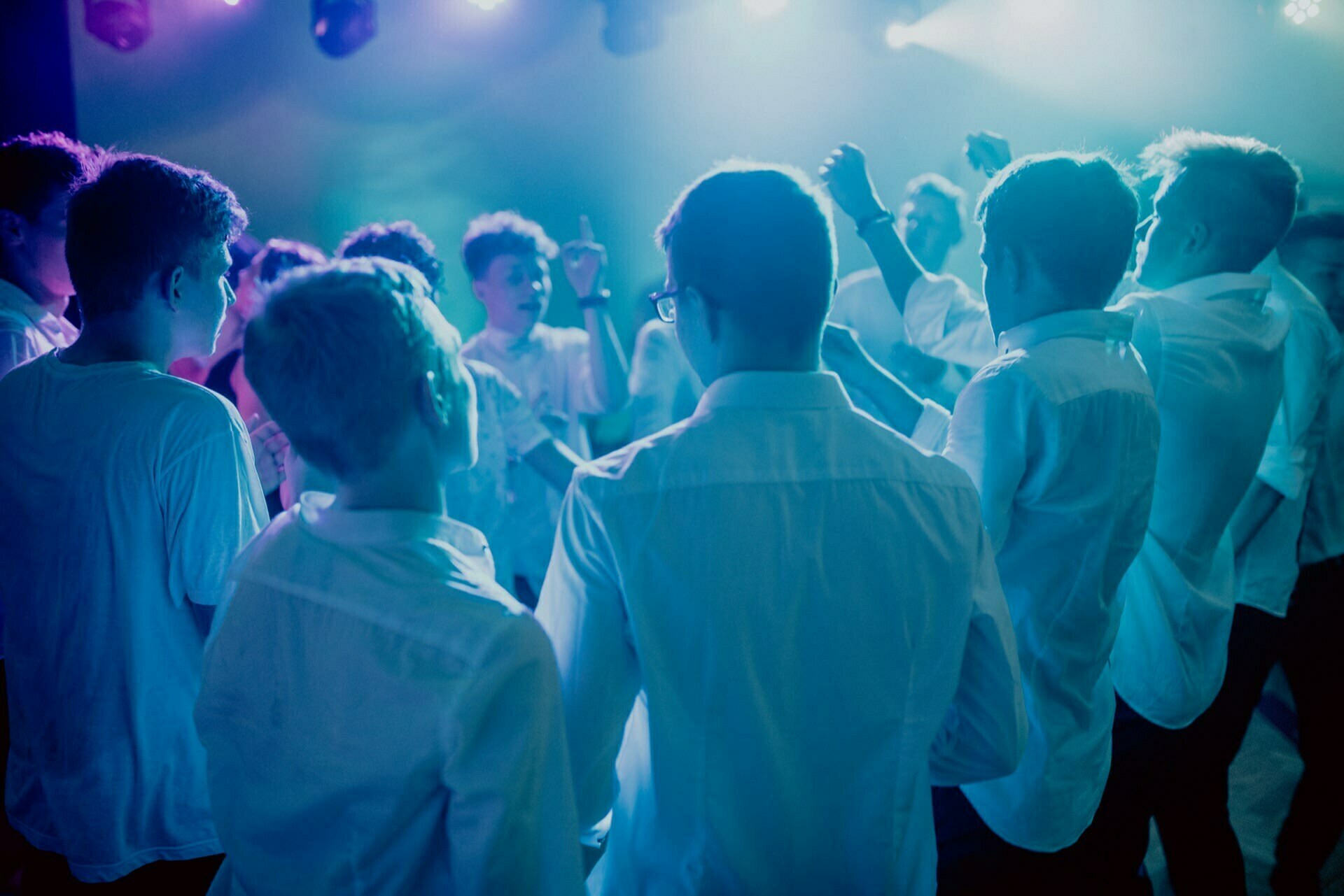 A group of people dressed in white shirts are dancing and celebrating in a dimly lit room with blue and purple lighting. They appear to be having a good time, raising their hands and moving energetically in circles. The atmosphere is lively and festive, perfect for an event photo essay.  