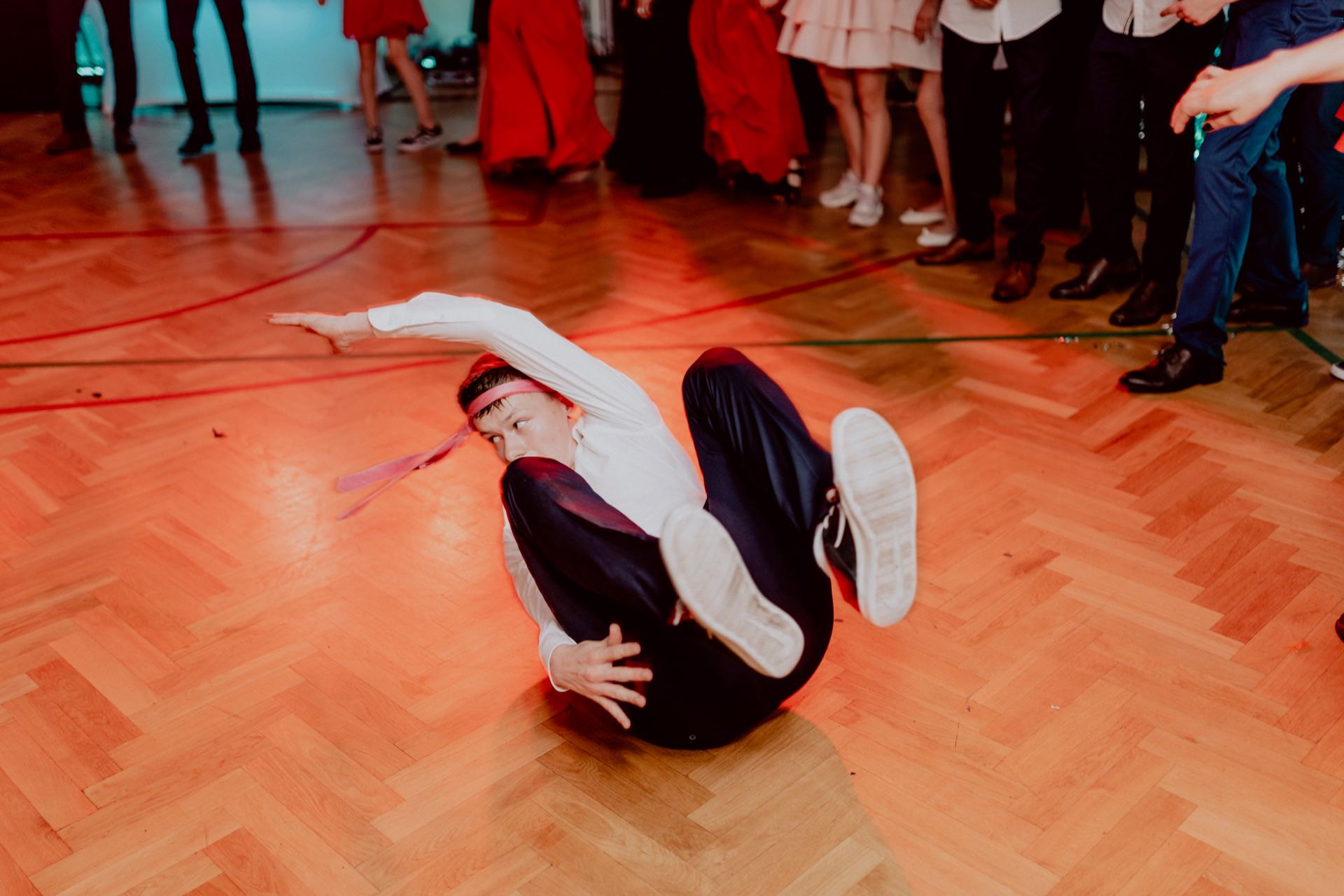 A person breakdancing on a wooden floor, surrounded by a crowd. The dancer is in mid-motion, lying on his back with his legs and arms raised. Red and blue lights illuminate the floor, capturing the essence of event photography. Other people in formal attire are visible in the background.   