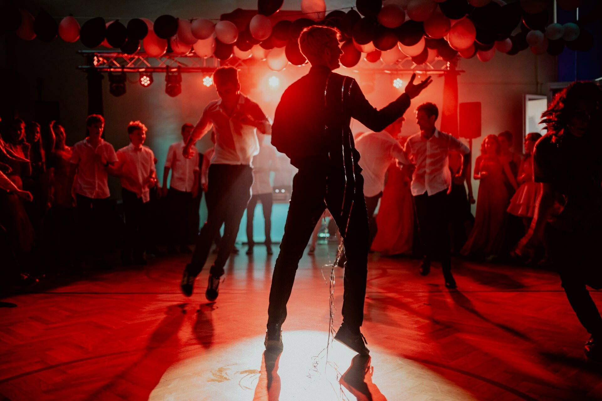 A person dances vigorously in the middle of a dimly lit room, illuminated by red and white lights. Other people dance and watch in the background as balloons hang from the ceiling, creating a festive atmosphere perfect for photo opportunities of the events. 