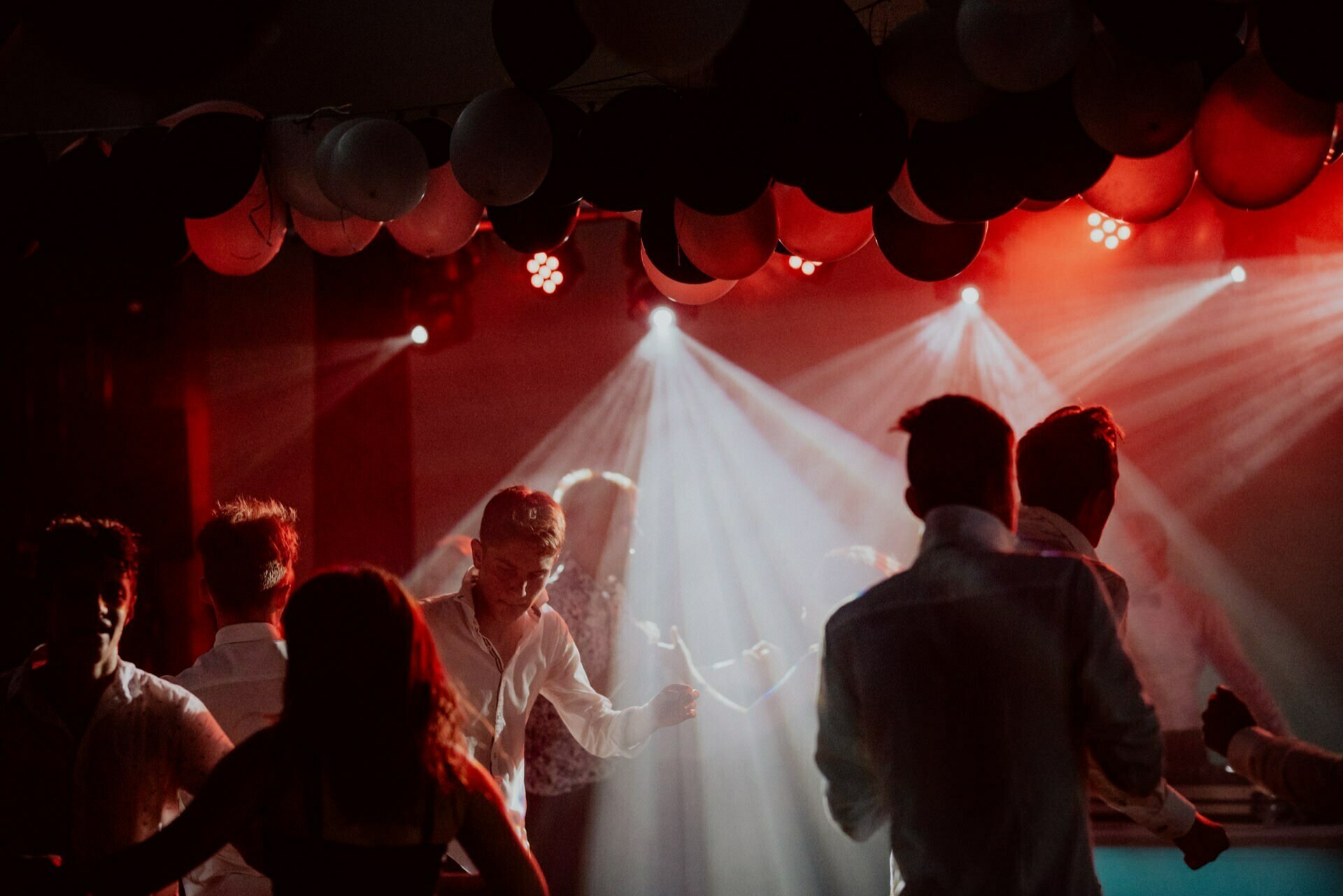 A vibrant party scene with people dancing under red and white stage lights. Black and white balloons hang from the ceiling, creating a festive atmosphere. The crowd depicts mostly dark silhouettes, with some people faintly illuminated by lights, perfectly capturing the essence of event photography.  