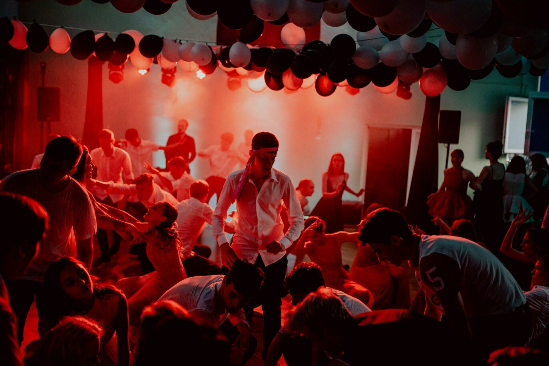 A crowd of people dance at a party under red and white balloon decorations. The room has an atmosphere of red light, and there seems to be a mist in the air, enhancing the atmosphere of a photographer at a middle school ball. People are having fun with energetic dance moves.  