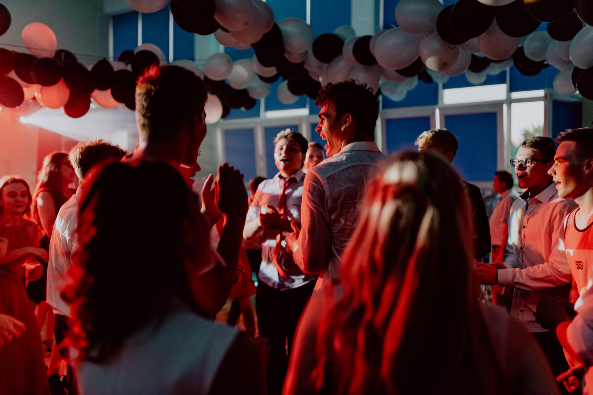 A lively group of people dance in a room decorated with black and white balloons hanging from the ceiling. The lighting is mostly red, creating a lively atmosphere, beautifully captured by the event photographer Warsaw. It looks like everyone is having a good time, talking and dancing.  