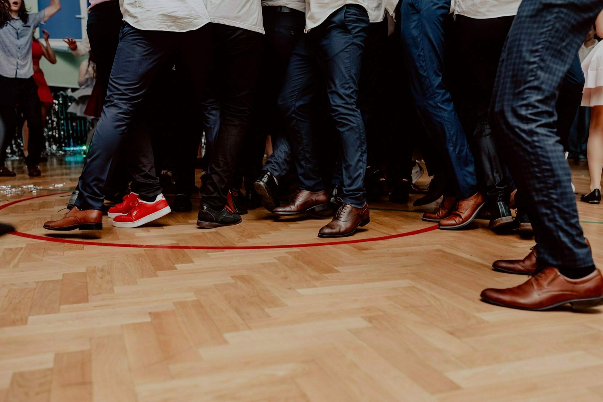 A group of people standing close together in a circle, viewed from the knees down, is captured in this event photograph. The bright wooden dance floor highlights their dark pants and various styles of formal shoes, except for one pair of bright red sneakers that stand out. 