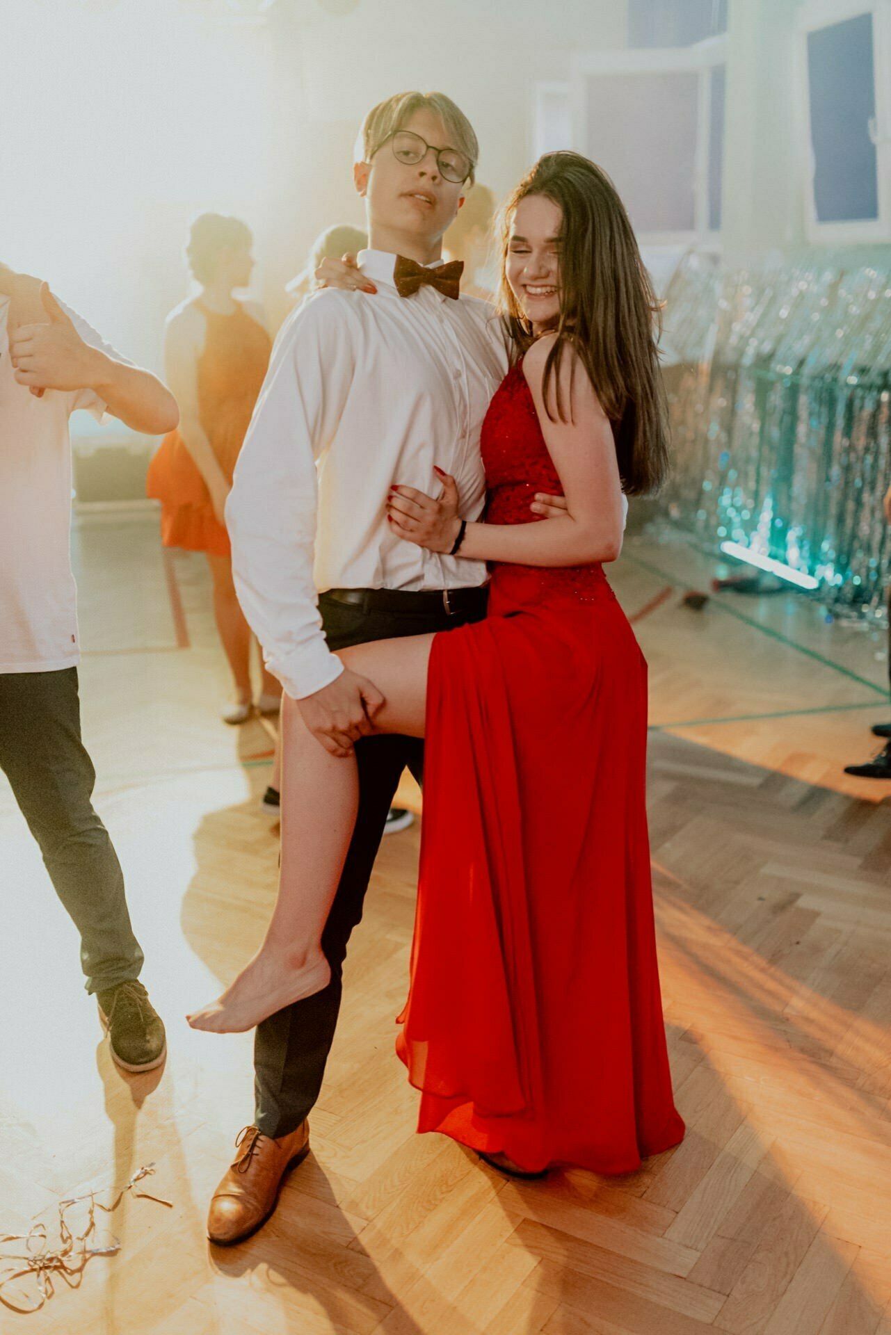 A young man in a white shirt and black bow tie is holding a young woman in a red dress, with one leg raised, barefoot. They are seated on a wooden dance floor with other people and decorations in the background, exuding a lively, festive atmosphere - a perfect scene for event photography. 