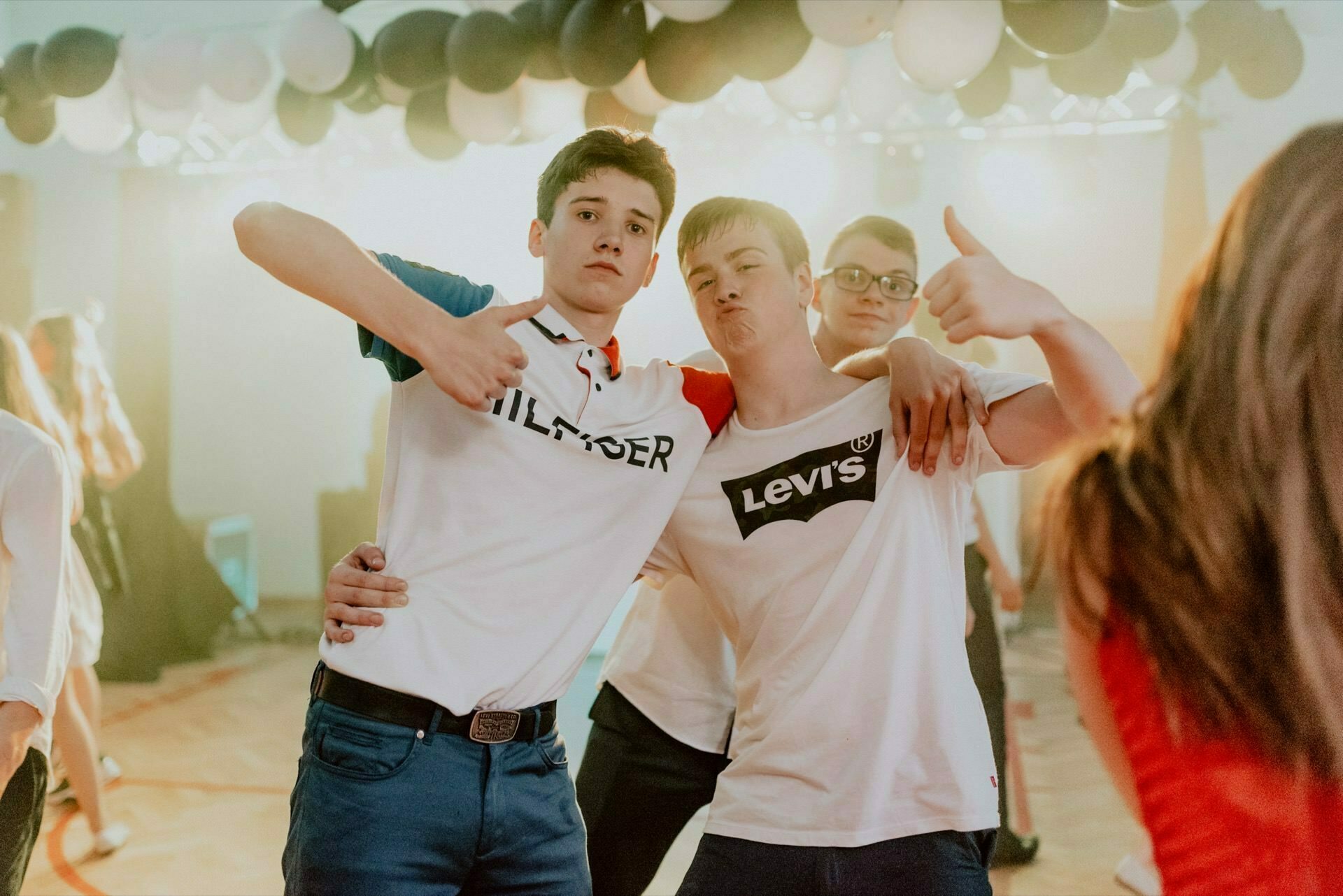 Three young men pose for a group photo at a social event. Two of them are standing close together, embracing each other's arms, showing thumbs up, while the third is standing slightly behind them, also showing thumbs up. This classic event photo shows balloons decorating the background.  