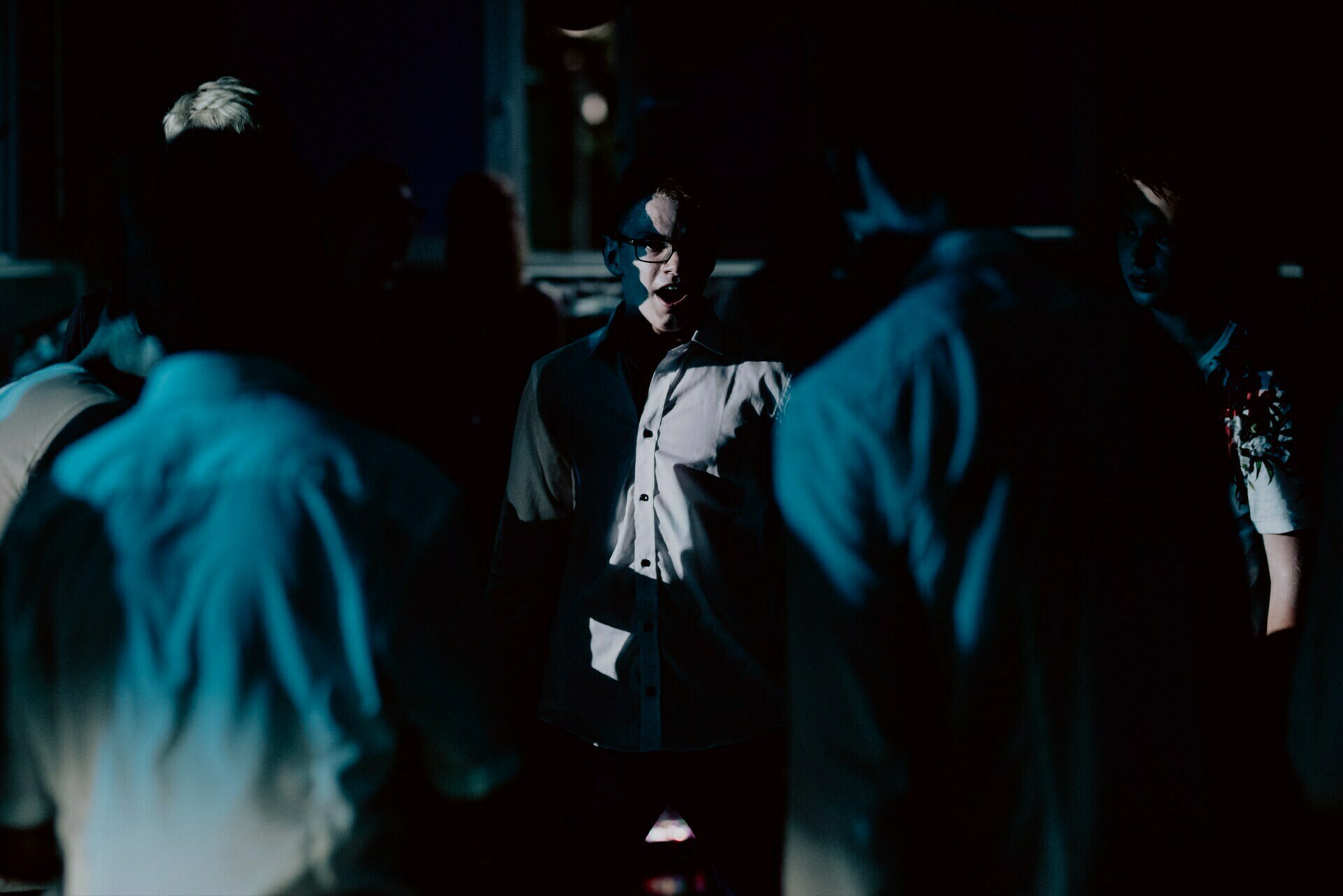 A person stands in the middle of a dimly lit room, facing the camera. The lighting casts dramatic shadows, partially illuminating some people in the surrounding crowd while leaving others in the dark, creating a tense atmosphere reminiscent of an intense photo-reportage of events. 