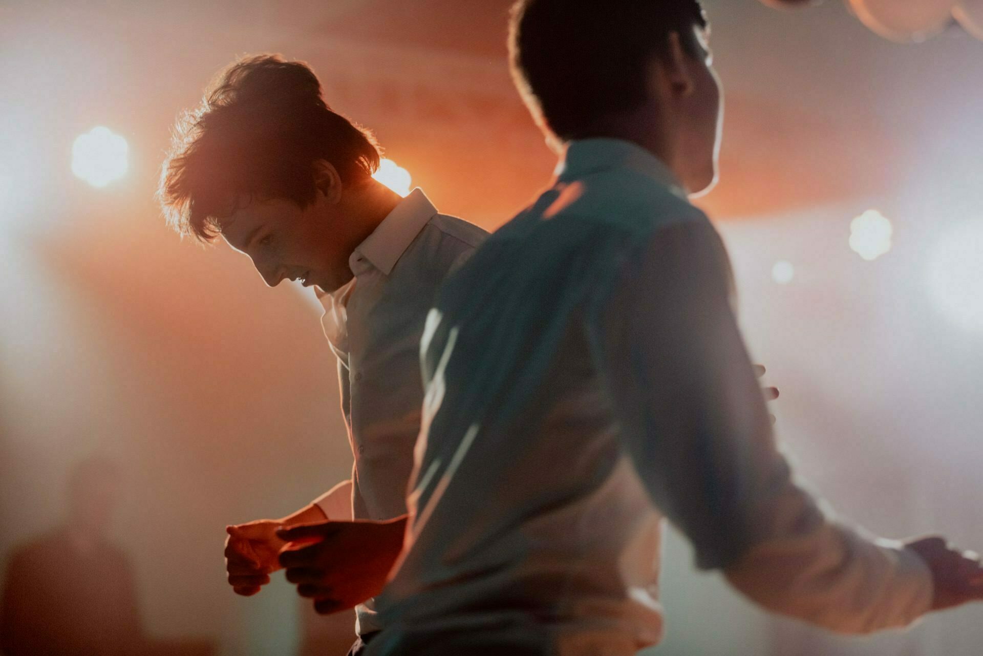 Two young men dressed in white shirts dance enthusiastically in warm light. The background is blurred with bright lights and faint silhouettes, creating an energetic atmosphere. Their movements convey joy and vitality, which was perfectly captured by the event photographer Warsaw for the event photo shoot.  