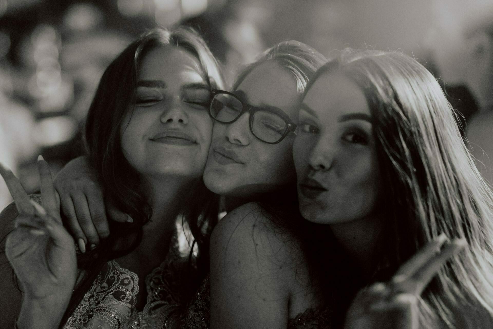 Black and white photo of three young women. They are smiling and making peace signs with their fingers. The photo gives the impression of a photo-reportage of events, where the blurred background shows other people and lights, enhancing the festive atmosphere.  