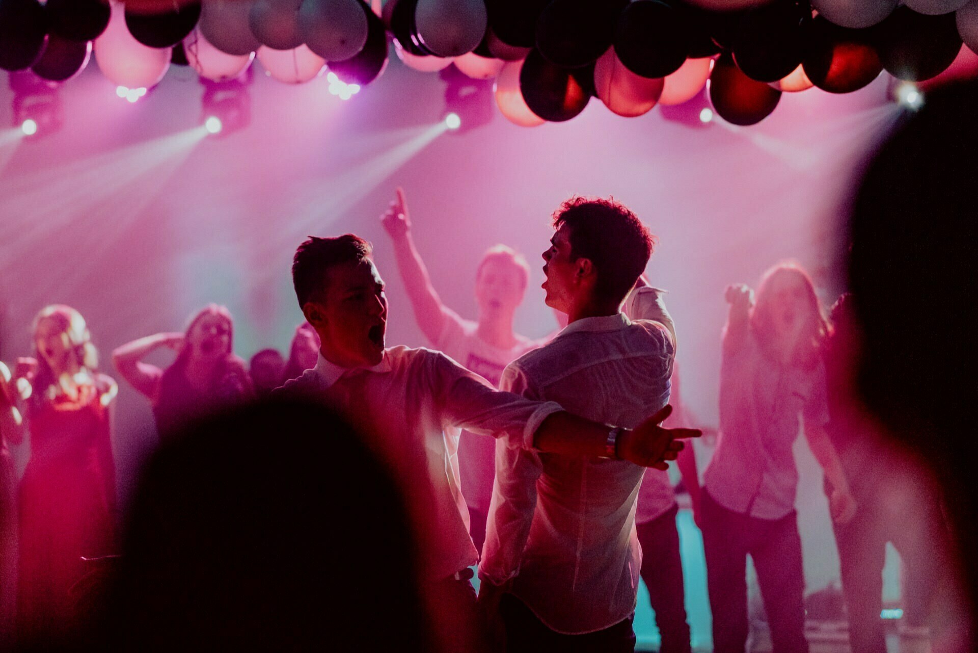 Two people dance energetically under hanging balloons and bright lights during a lively party. They are surrounded by a crowd that is also dancing and having a good time. The vibrant, colorful atmosphere is full of movement and excitement, which is perfectly captured in the event photo report by event photographer Warsaw.  