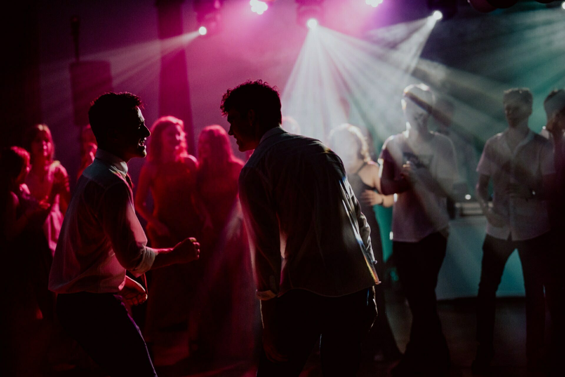 A dimly lit club scene with vibrant pink and blue lights illuminating energetic dancing people. The photo captures a lively atmosphere with people enjoying the music, some holding drinks and expressing various expressions of joy and excitement - perfect for an event photography show. 