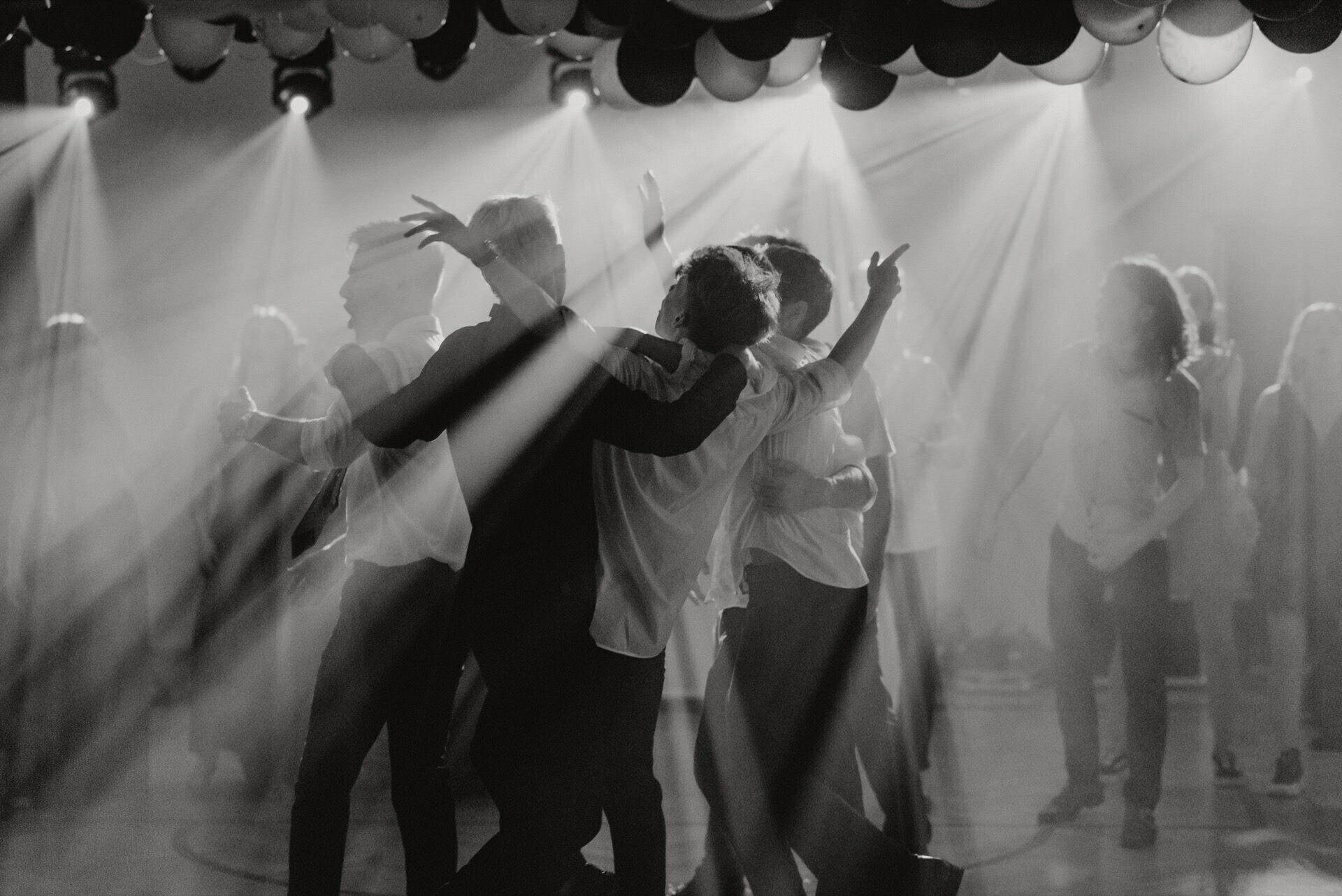 Black and white photo of a group of people dancing energetically under a ceiling decorated with balloons. Streams of light and smoke create an atmospheric effect, giving a festive and lively atmosphere. Other participants can be seen in the background, capturing the essence of event photography.  