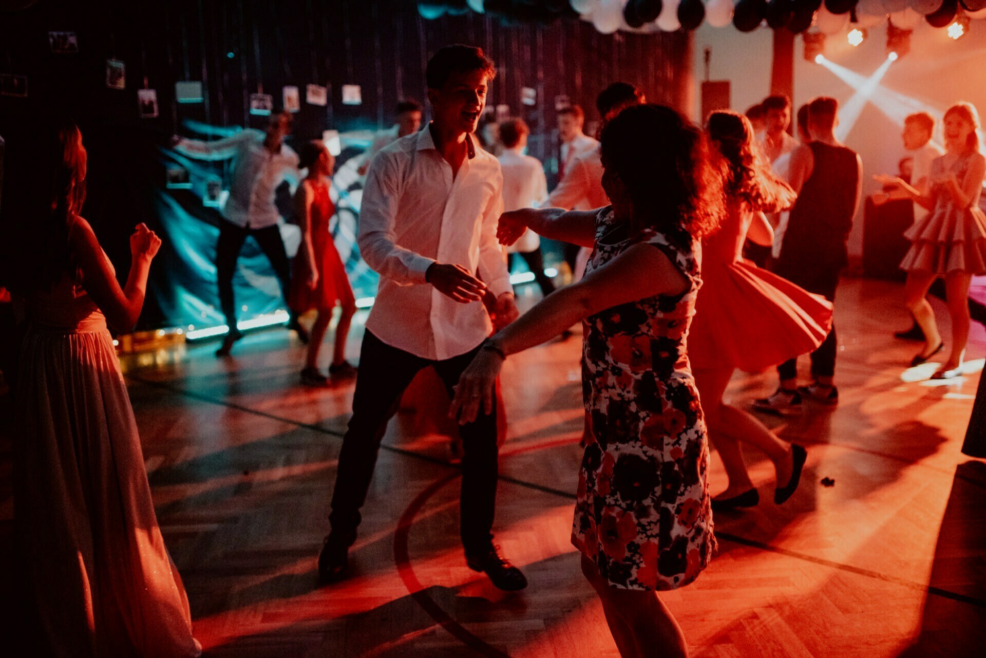 People are dancing at a dimly lit party. A man in a white shirt and a woman in a flowery dress can be seen dancing in the foreground. Bright lights and decorations, including balloons and photos on the wall, create a festive atmosphere - perfectly captured by event photographer Warsaw during the memorable event photo shoot.  