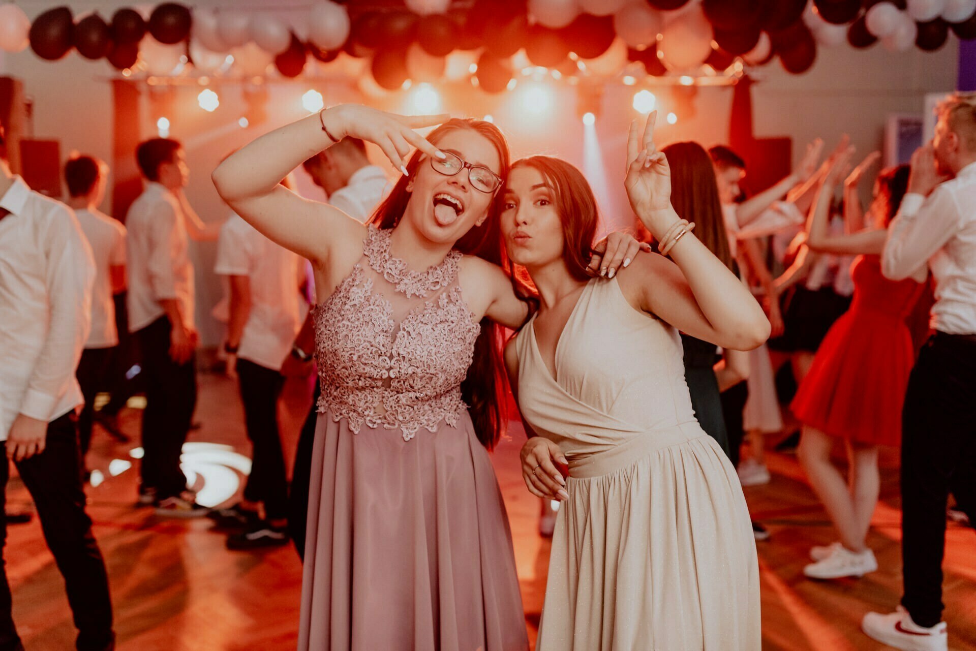 Two young women in elegant dresses assume playful poses on the dance floor during a lively party. One is dressed in a lavender dress and sticks out her tongue, while the other in a white dress makes a peace sign. The background is filled with dancing people and festive balloons in the warm light - a perfect photo-op of the events.  