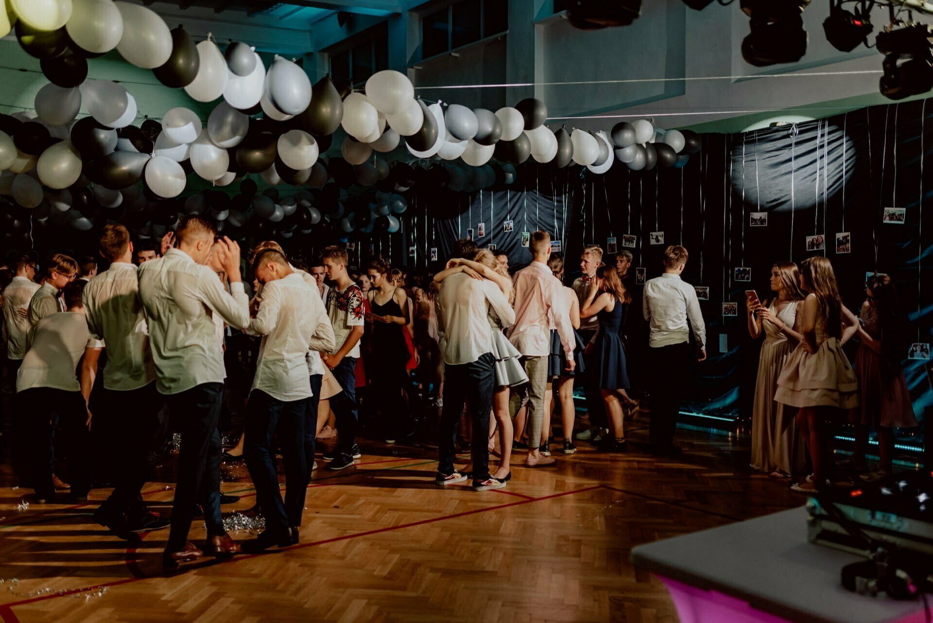 A group of people are attending a dance party in a decorated room with black and white balloons on the ceiling. Some dance closely in pairs, while others stand and talk. The backdrop is decorated with hanging photos, creating an intimate atmosphere ideal for photo coverage of the events.  