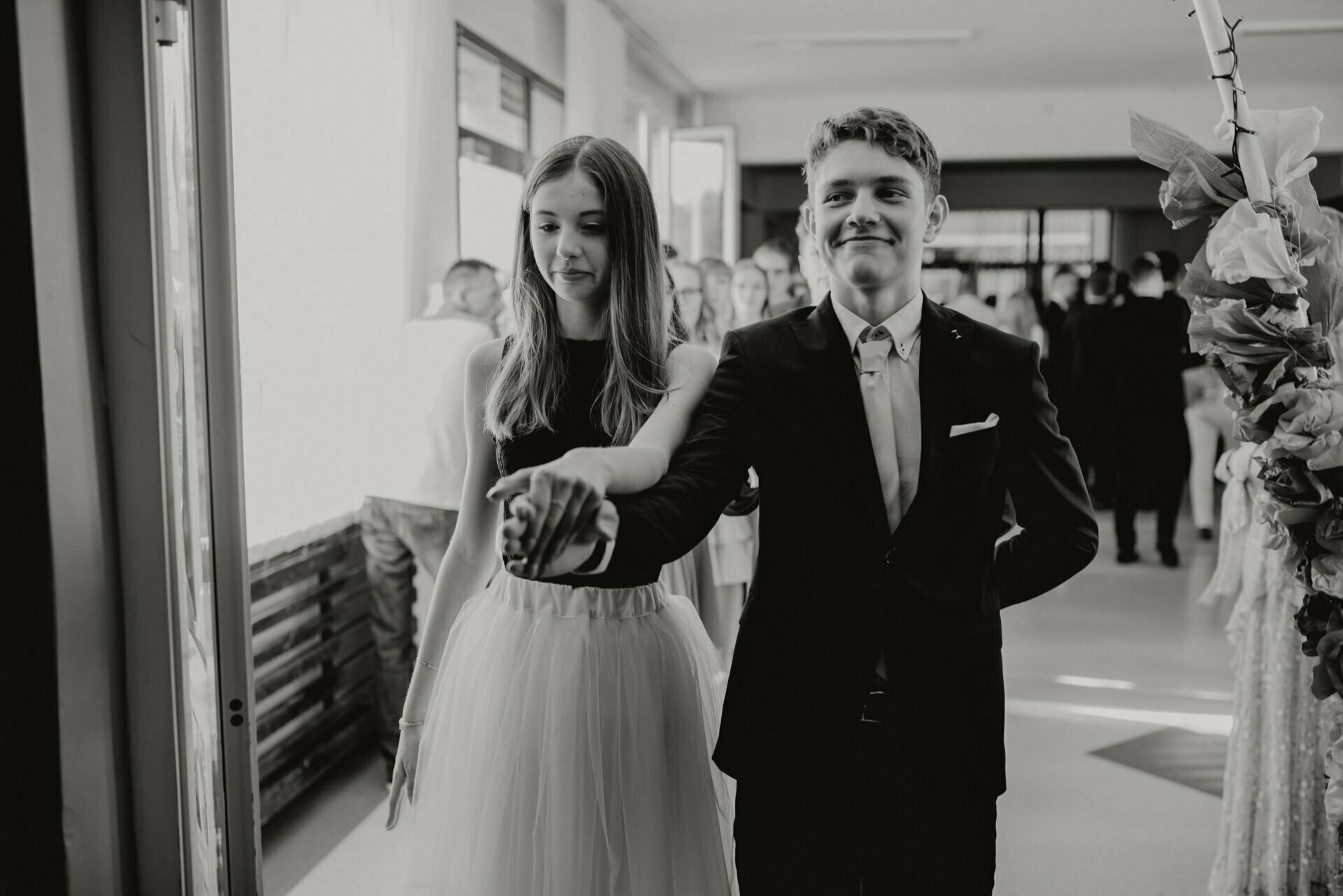 A young couple holding hands and walking through a doorway, dressed in formal attire. The young man is dressed in a suit and bow tie, while the young woman is wearing a sleeveless dress and tulle skirt. Captured by the event photographer warsaw, they appear to be heading towards the event, with several people visible in the background.  