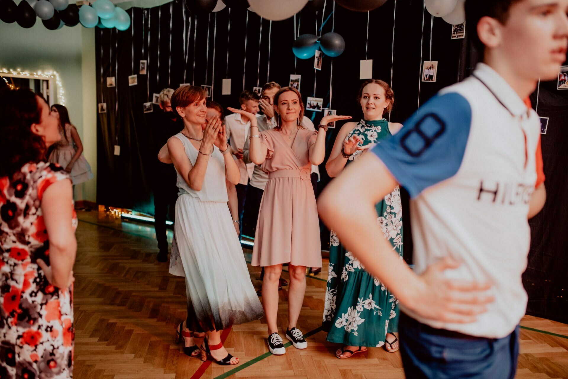 People dressed in semi-formal attire are enjoying themselves at a party in a decorated venue with balloons and photos on the walls. In the center of the photo, several women are clapping and smiling, and a person in a navy blue and white shirt is partially visible in the foreground - perfect moments for event photography by any event photographer Warsaw. 