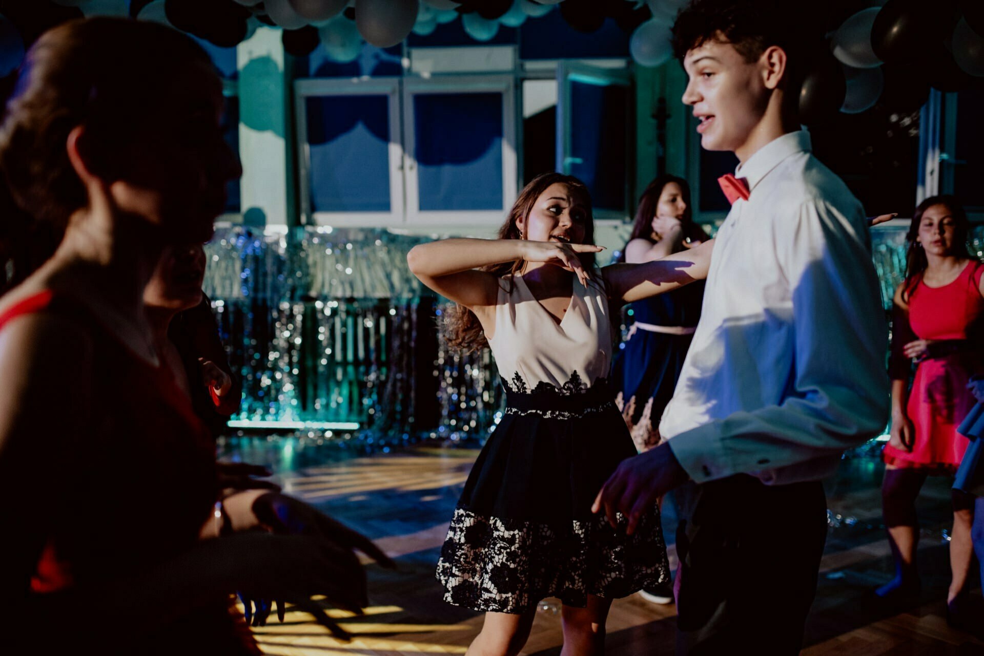 A group of young people dance energetically in a decorated indoor space. In the background are balloons and metallic streamers. A young woman in a white top and black skirt and a young man in a white shirt and red bow tie are perfectly visible, perfectly captured in this event photography.  