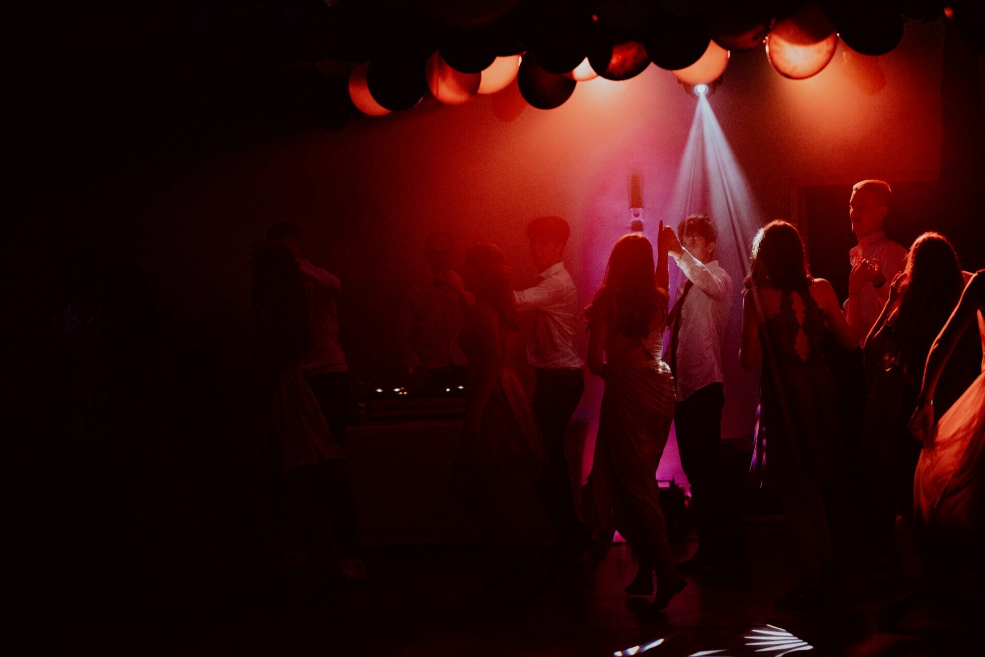 A dimly lit dance floor where people dance in red light. The ceiling is decorated with black and red balloons. A spotlight falls from above, illuminating the dancers. The atmosphere seems lively, and silhouettes of people enjoy the music - perfect for capturing event photography in Warsaw.   